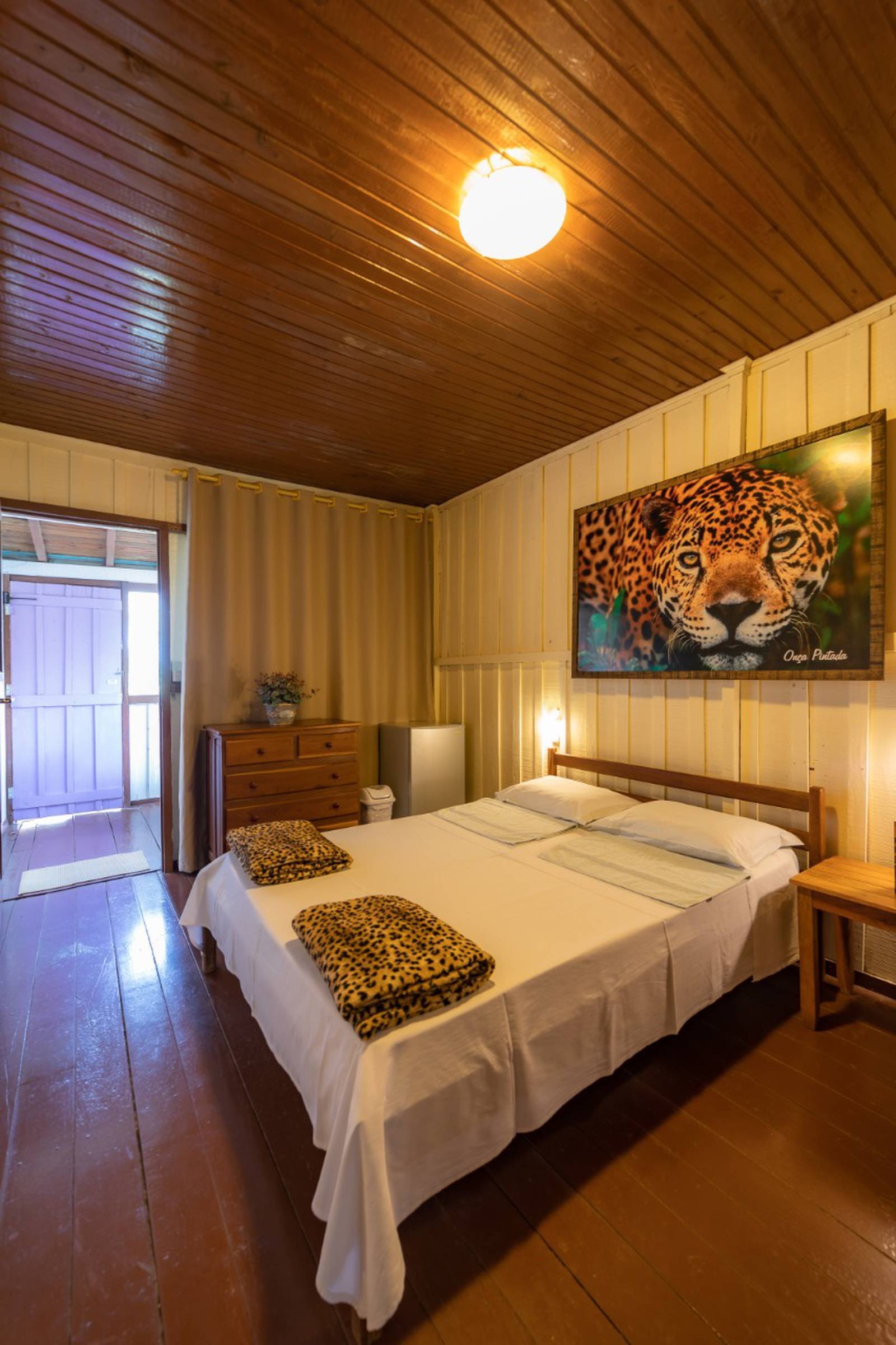 Cosy double room at a budget Pantanal tour lodge, featuring a warm wooden ceiling, leopard-print blankets on the bed, and a large jaguar portrait on the wall