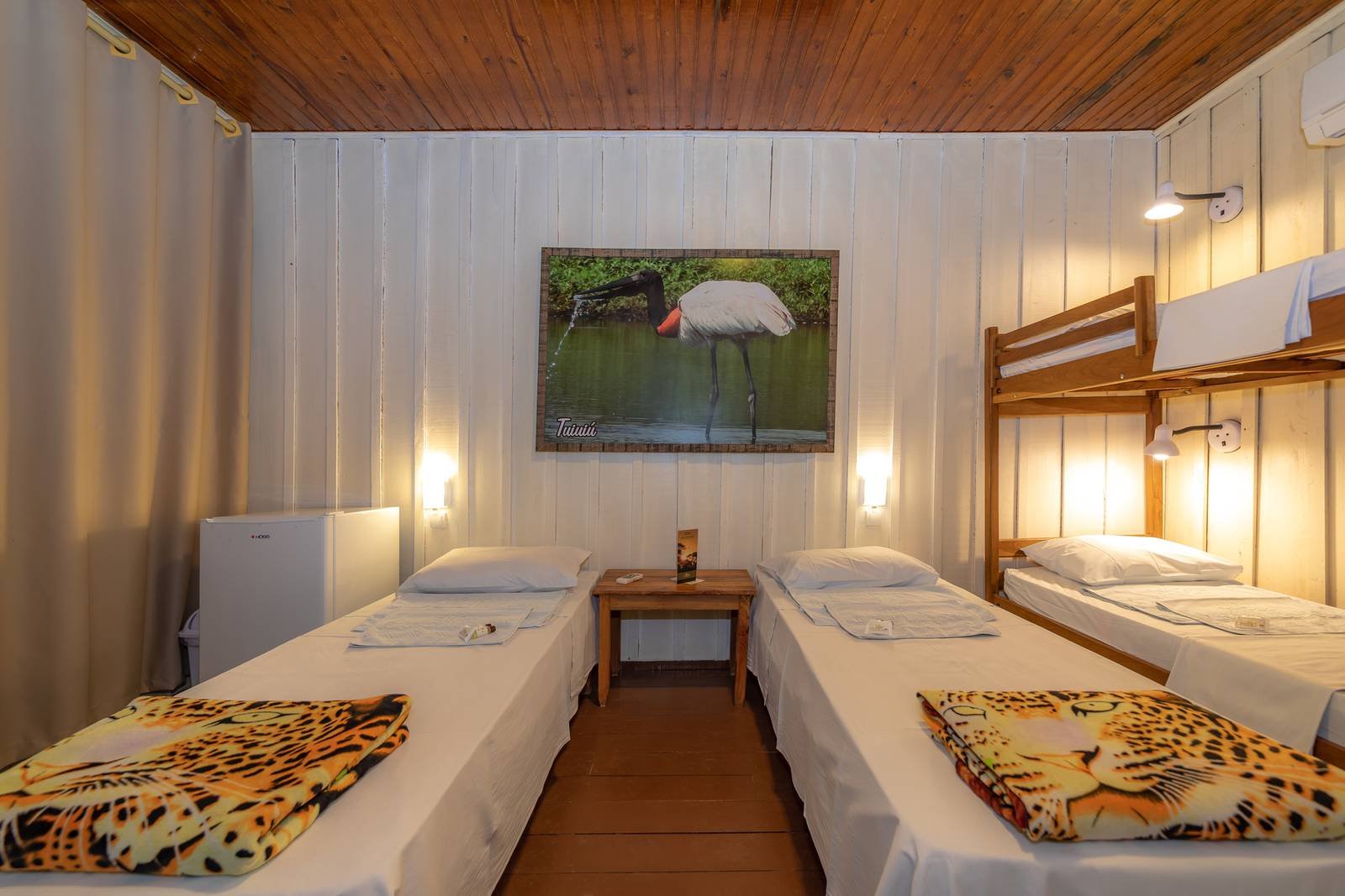 Cozy multi-bed lodge room on a budget pantanal tour, featuring two single beds with jaguar-print blankets, a wooden bunk bed, warm wall lighting, and a framed jabiru stork artwork above a small bedside table