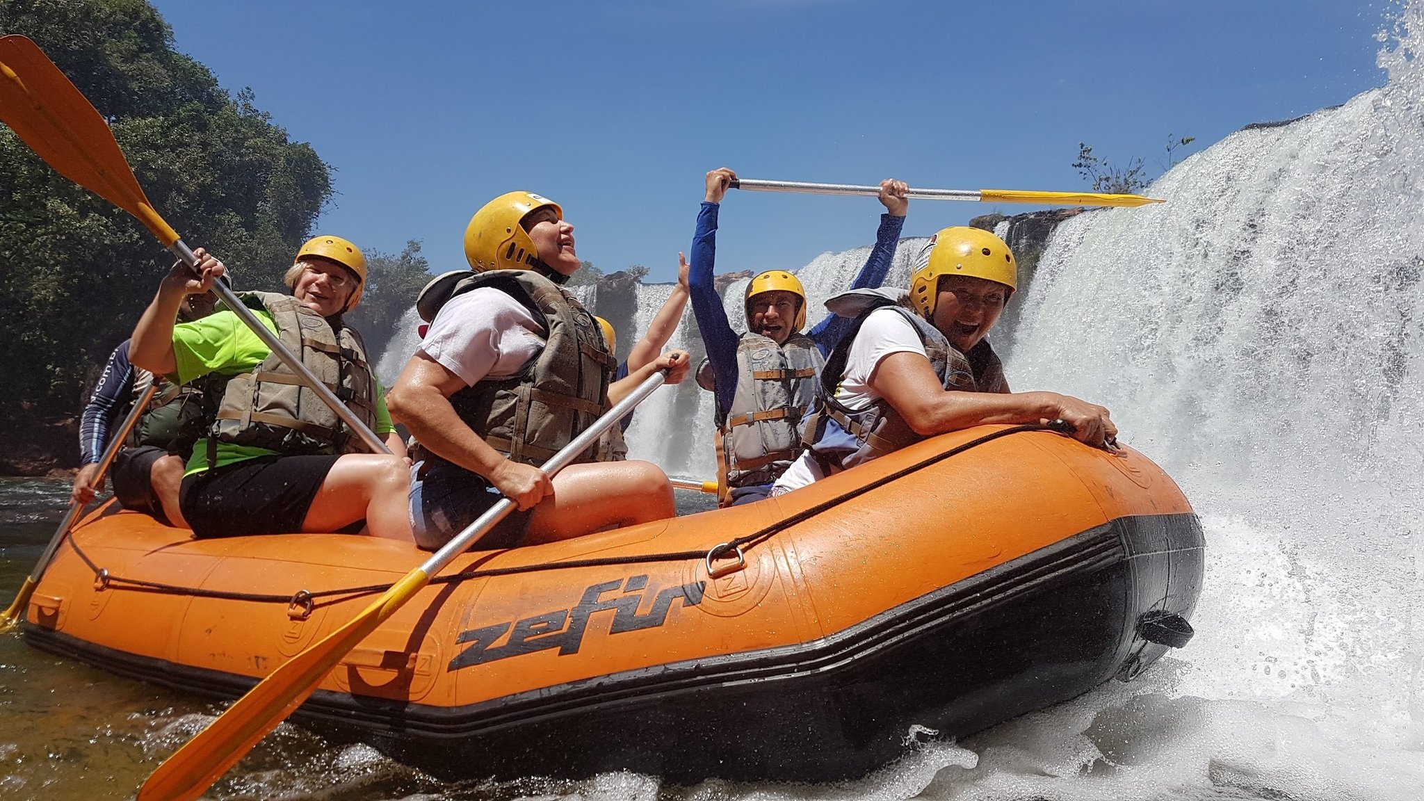 Cinco aventureiros com capacetes amarelos e coletes salva-vidas remam em um bote laranja ao lado de uma cachoeira imponente no Cerrado Brasil