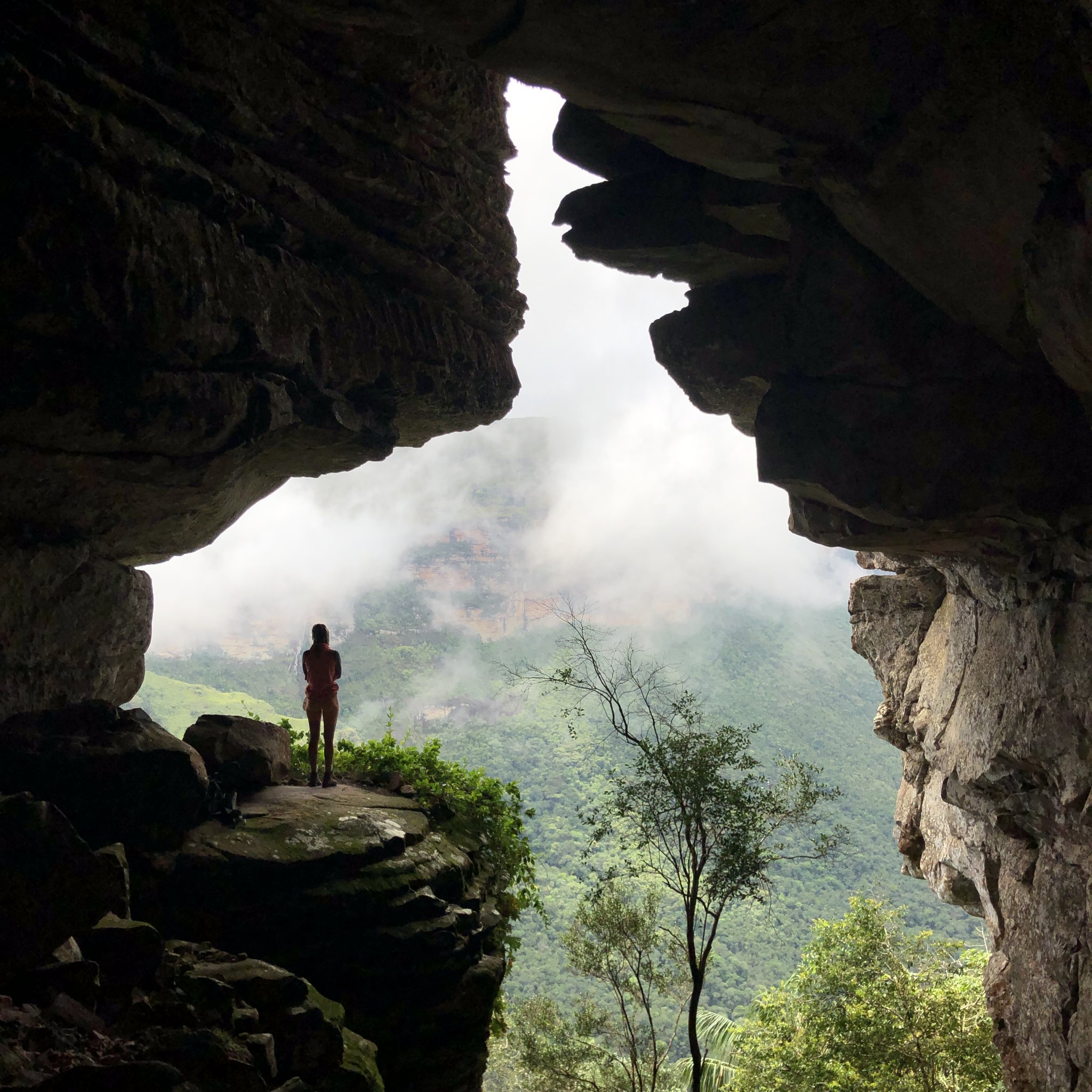 A hiker silhouetted inside a dramatic rock cave opening, gazing out over the cloud-wrapped forested mountains of Chapada Diamantina