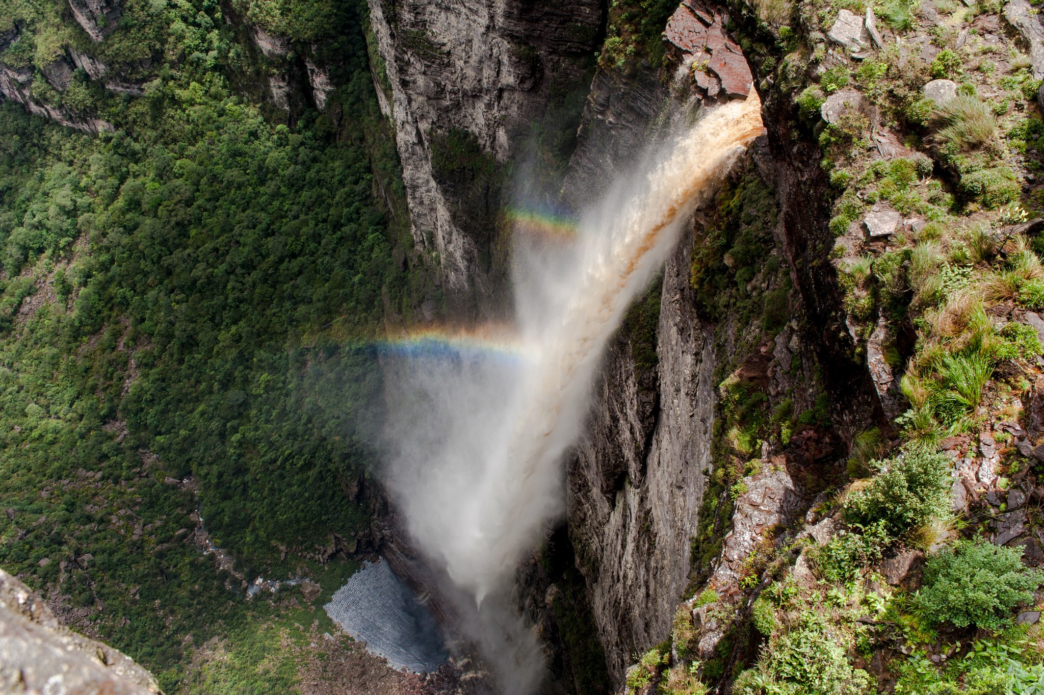Aerial view looking straight down at a powerful waterfall plunging through a narrow rocky canyon in Chapada Diamantina, with a vivid rainbow forming in the mist above the pool below