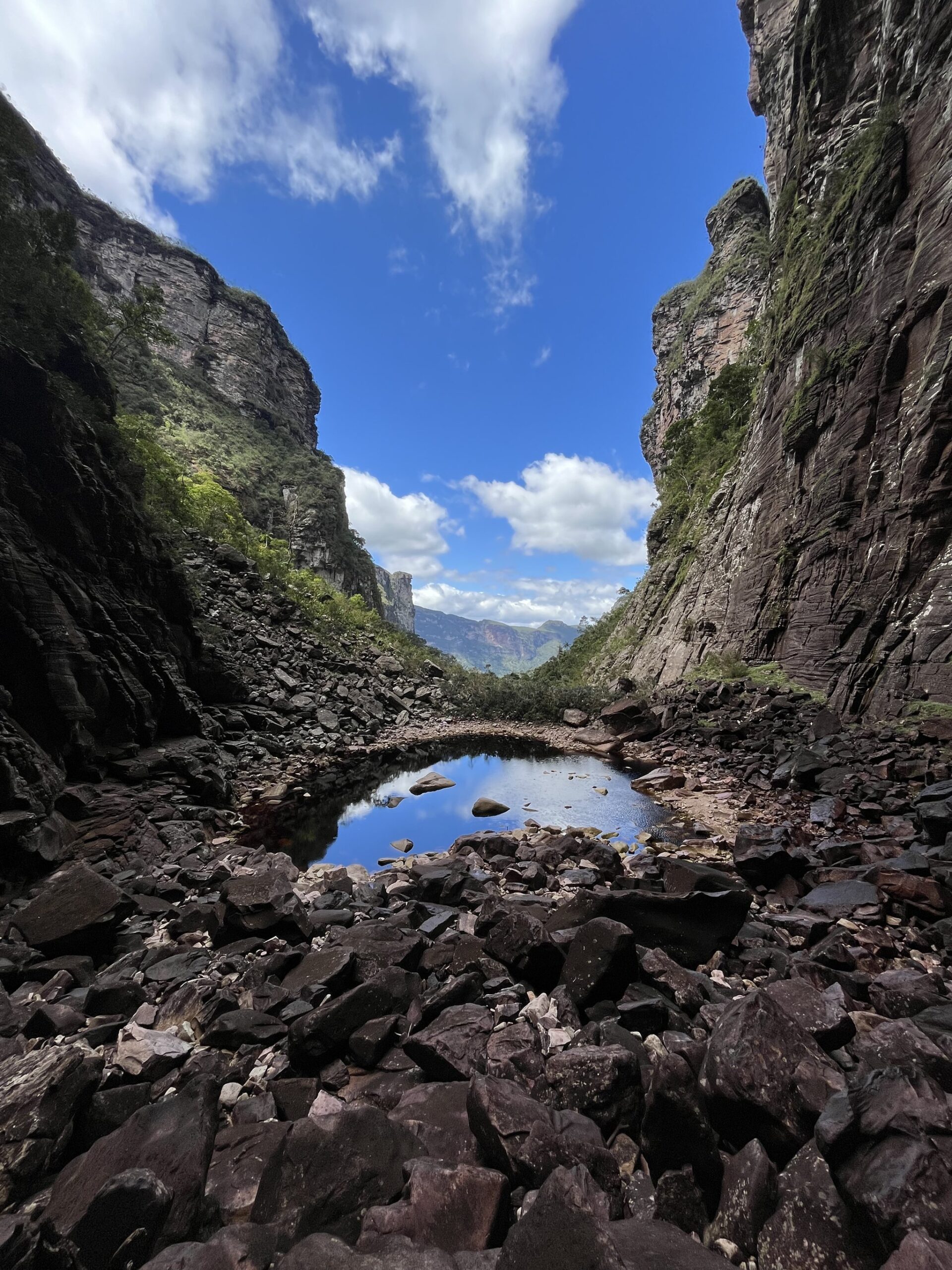 Sky caught in stone — a quiet pool between canyon walls. Photo: Lucas Neves A still rockpool mirrors the blue sky between towering sandstone canyon walls on the Chapada Diamantina trek, with scattered boulders leading toward open mountains in the distance
