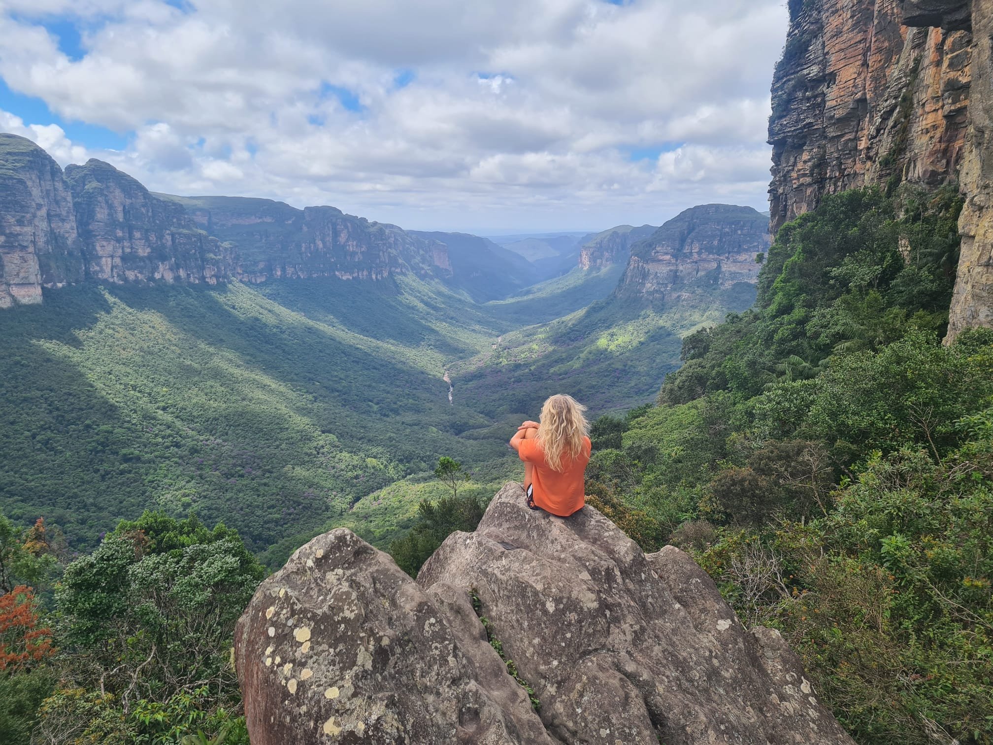 Perched on the edge, the whole valley yours to absorb. A hiker in an orange jacket sits on a rocky ledge taking in the Chapada Diamantina trek panorama, with a vast green valley flanked by towering sandstone canyon walls stretching to the horizon