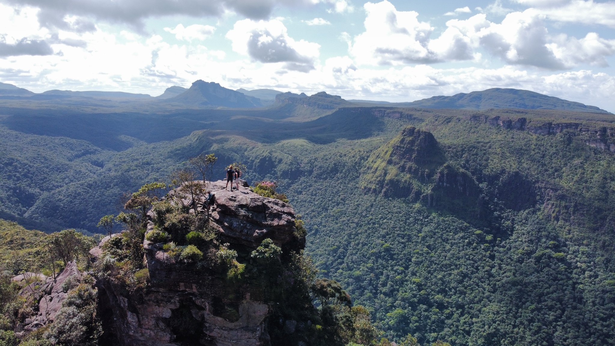 Summit reached — the whole canyon yours. Photo: Alex Hubin Two hikers standing on an exposed rock summit during a Chapada Diamantina trek, overlooking a sweeping forested canyon and distant sandstone plateaus under a partly cloudy sky