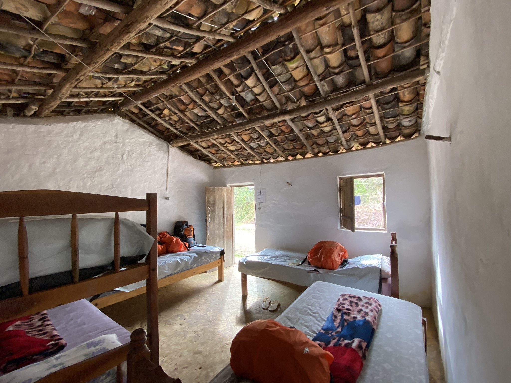 Clay tiles overhead, beds ready for weary trekkers — a local family home in the heart of the valley. Simple shared bedroom in a Vale do Pati homestay, with wooden bunk beds, single beds loaded with trekkers' orange dry bags and backpacks, white-washed walls and an exposed terracotta-tile roof on timber beams — typical Chapada Diamantina trek accommodation