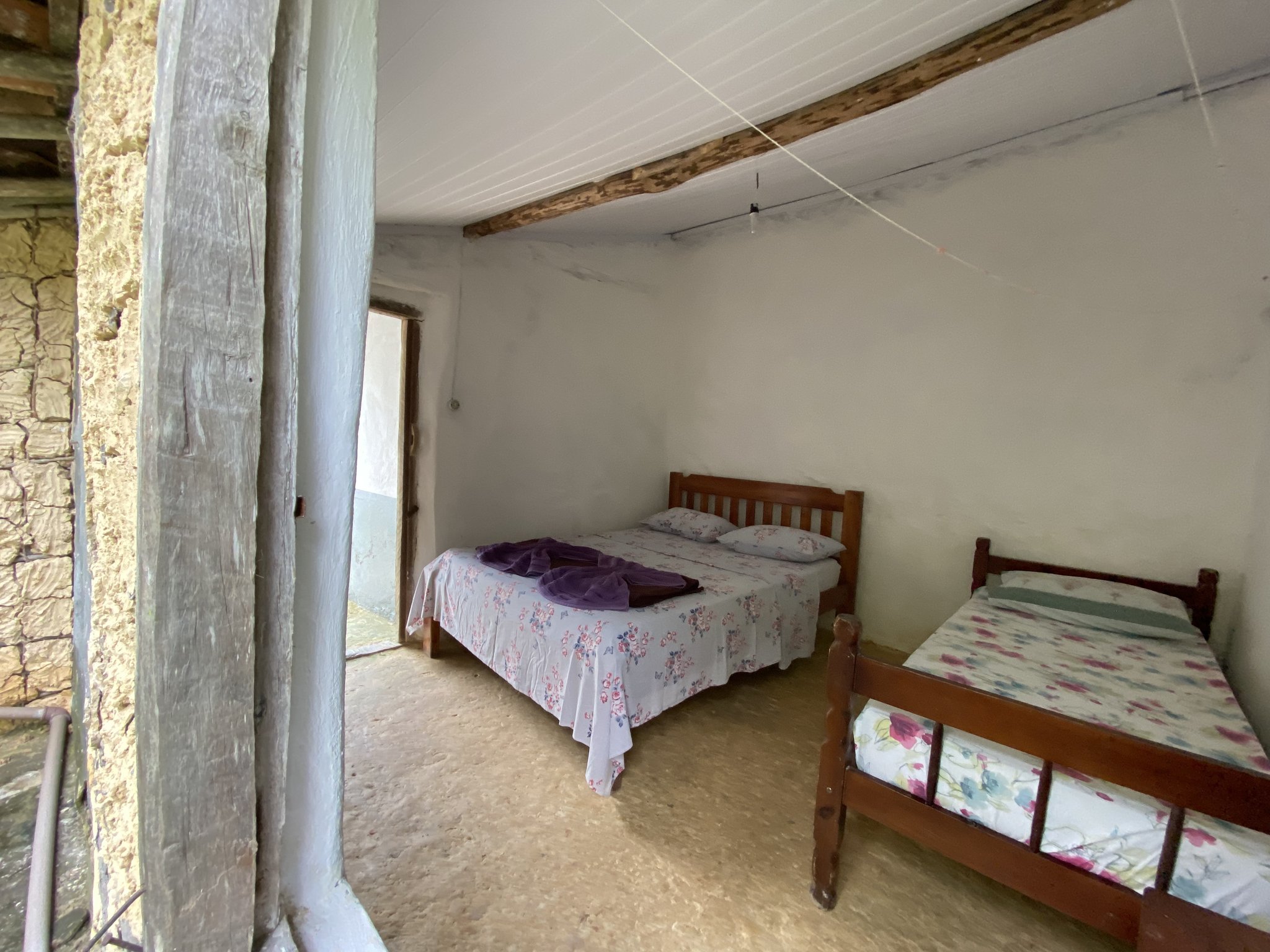 Rustic beds, stone walls, and a door open to the mountains — comfort enough after a day on the trail. Simple bedroom in a local family homestay on the Chapada Diamantina trek, with two wooden-framed beds dressed in floral bedding, bare stone walls and exposed ceiling beams