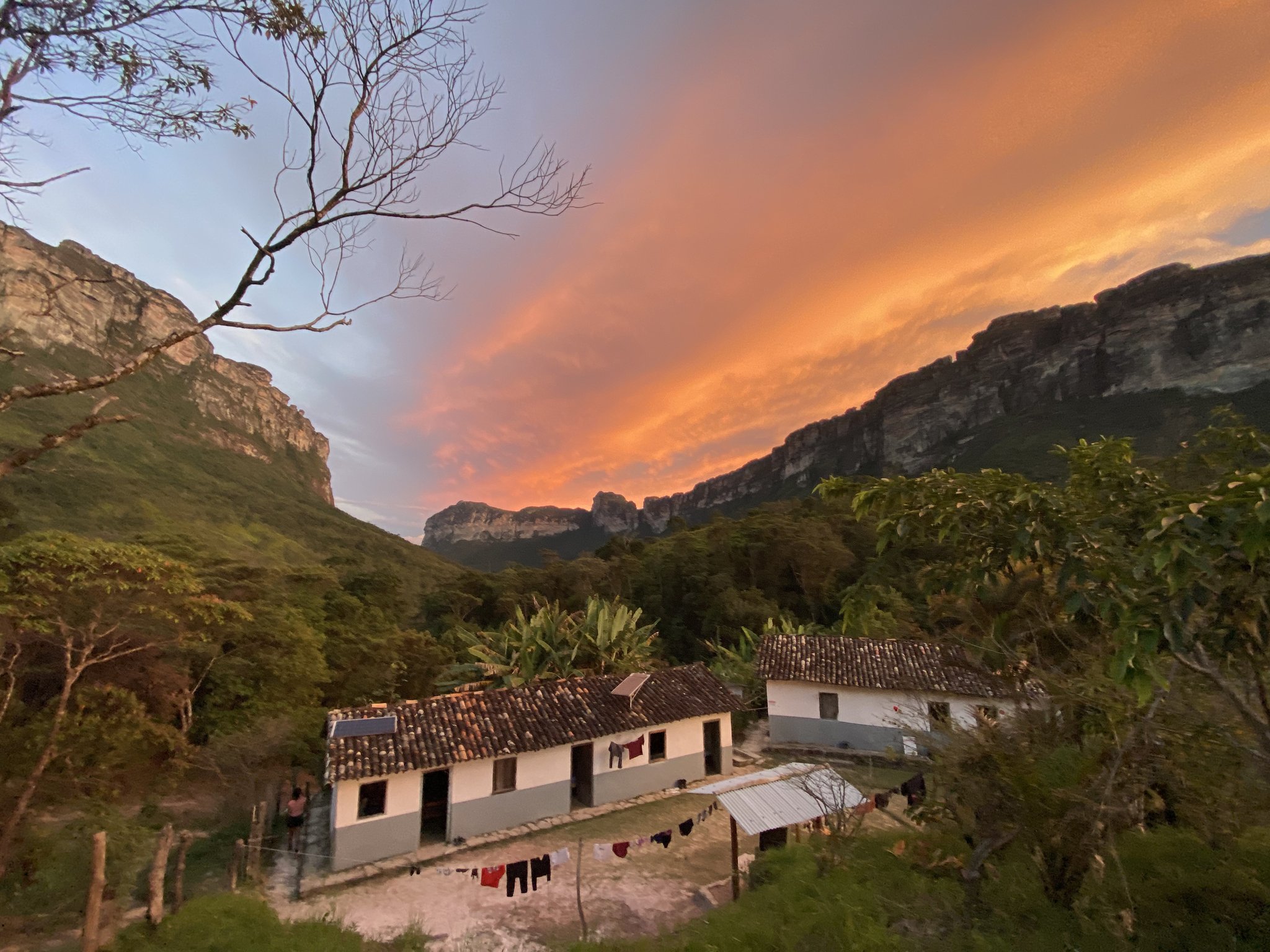 Laundry swaying, cliffs glowing — an evening at a local family home inside the valley. Rustic whitewashed homestay buildings with terracotta roofs nestled in a steep-walled valley on the Chapada Diamantina trek, laundry hanging on a line as a fiery orange sunset lights up dramatic sandstone cliffs above
