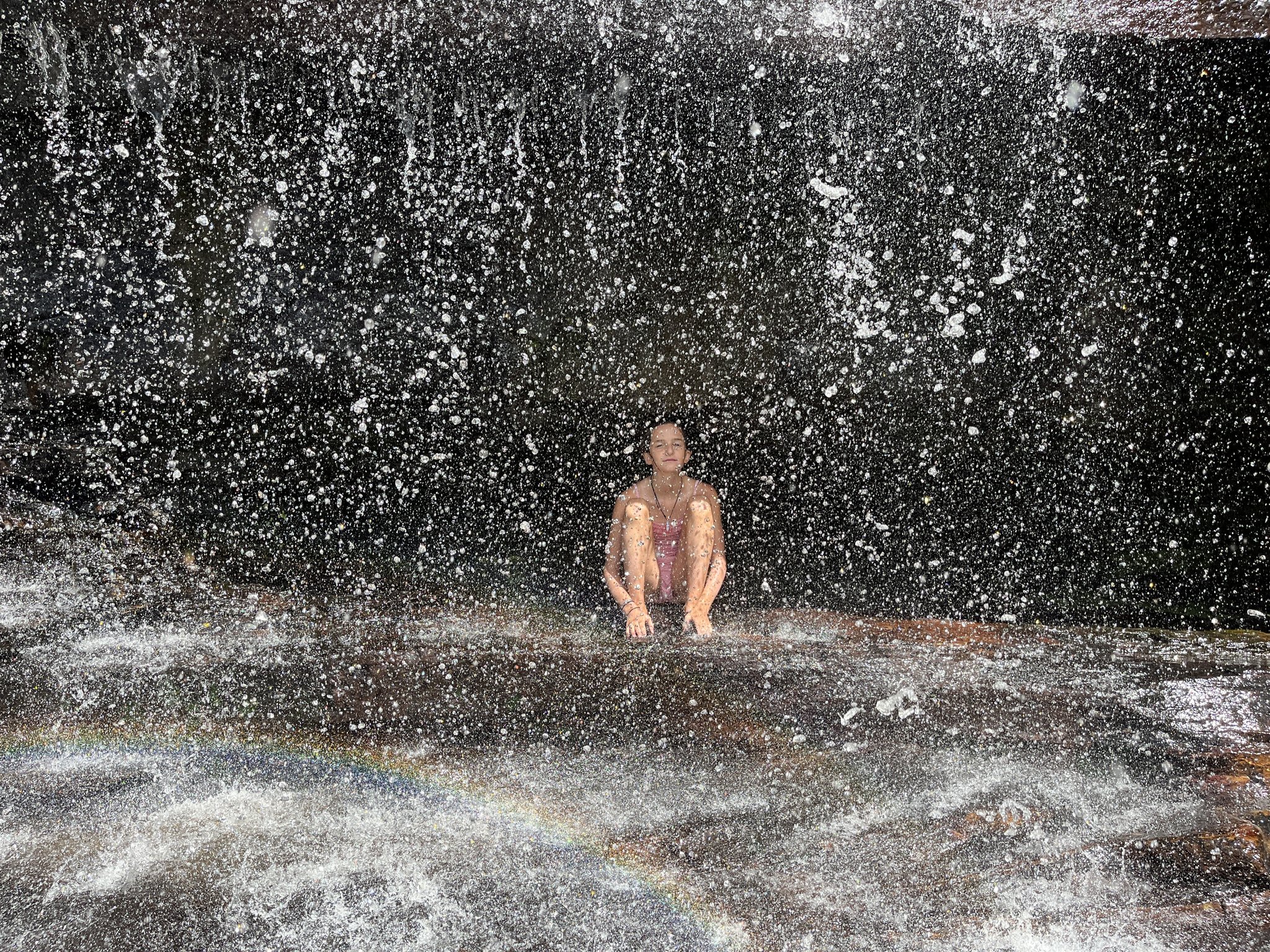 A traveler sits serenely on a wet rock ledge beneath cascading waterfall spray during a Chapada Diamantina trek, a tiny rainbow shimmering in the mist at her feet