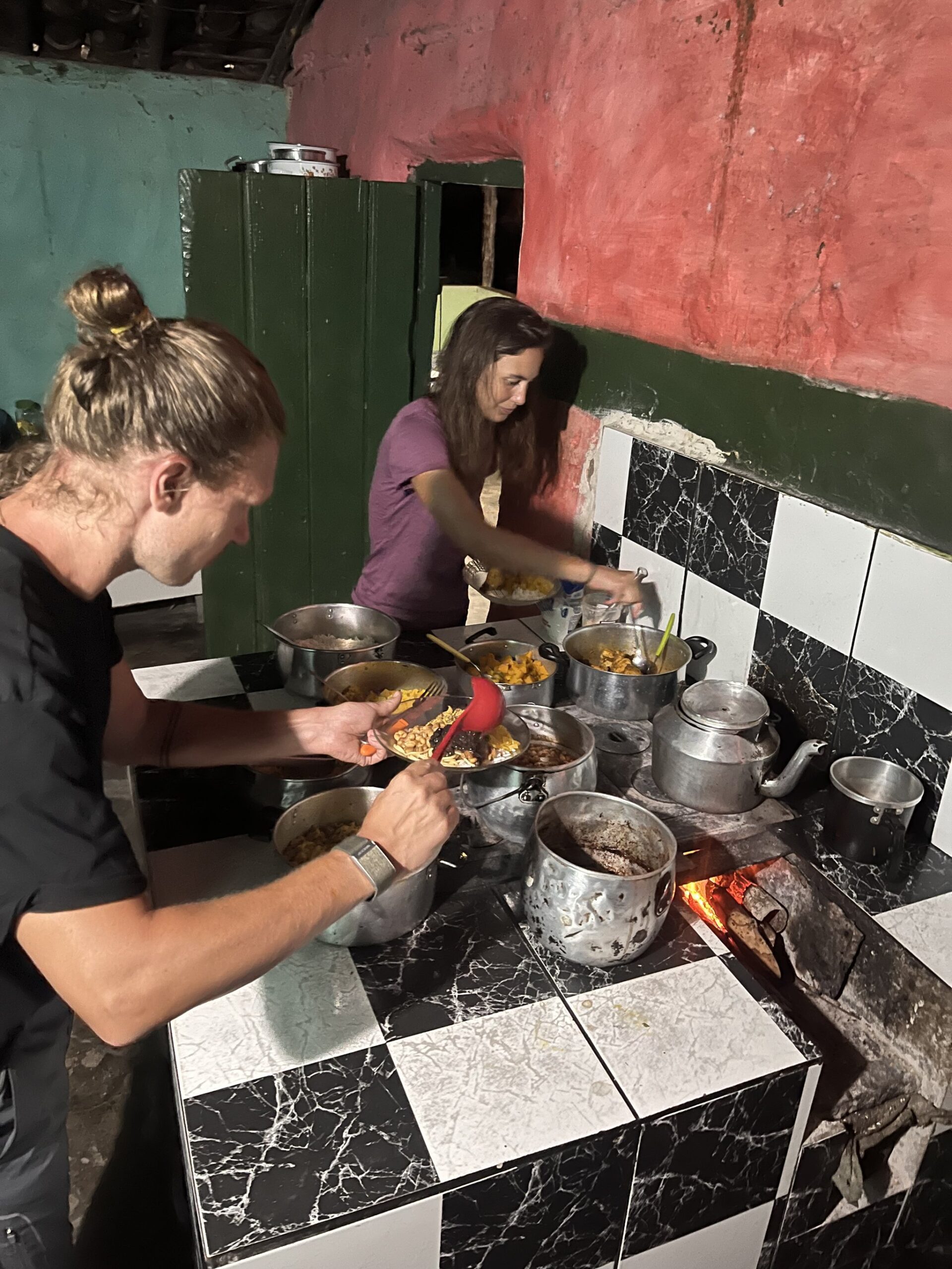 Real fire, real food — scooping up a home-cooked dinner after a long day on the trail. Photo: Lucas Neves Two travelers serving themselves from multiple pots on a wood-fired stove during a Chapada Diamantina trek homestay dinner, in a rustic kitchen with black-and-white tiled counters and colourful painted walls