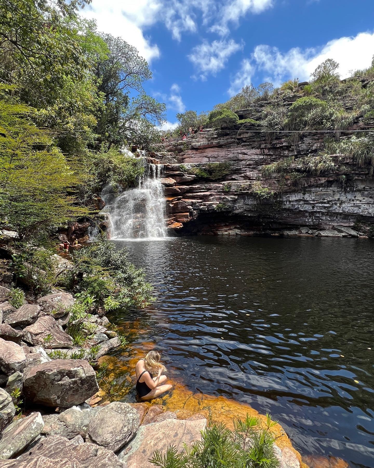 A traveller sits at the edge of a clear natural pool fed by a tiered waterfall cascading over layered sandstone cliffs on a Chapada Diamantina trip, surrounded by lush green vegetation under a bright blue sky
