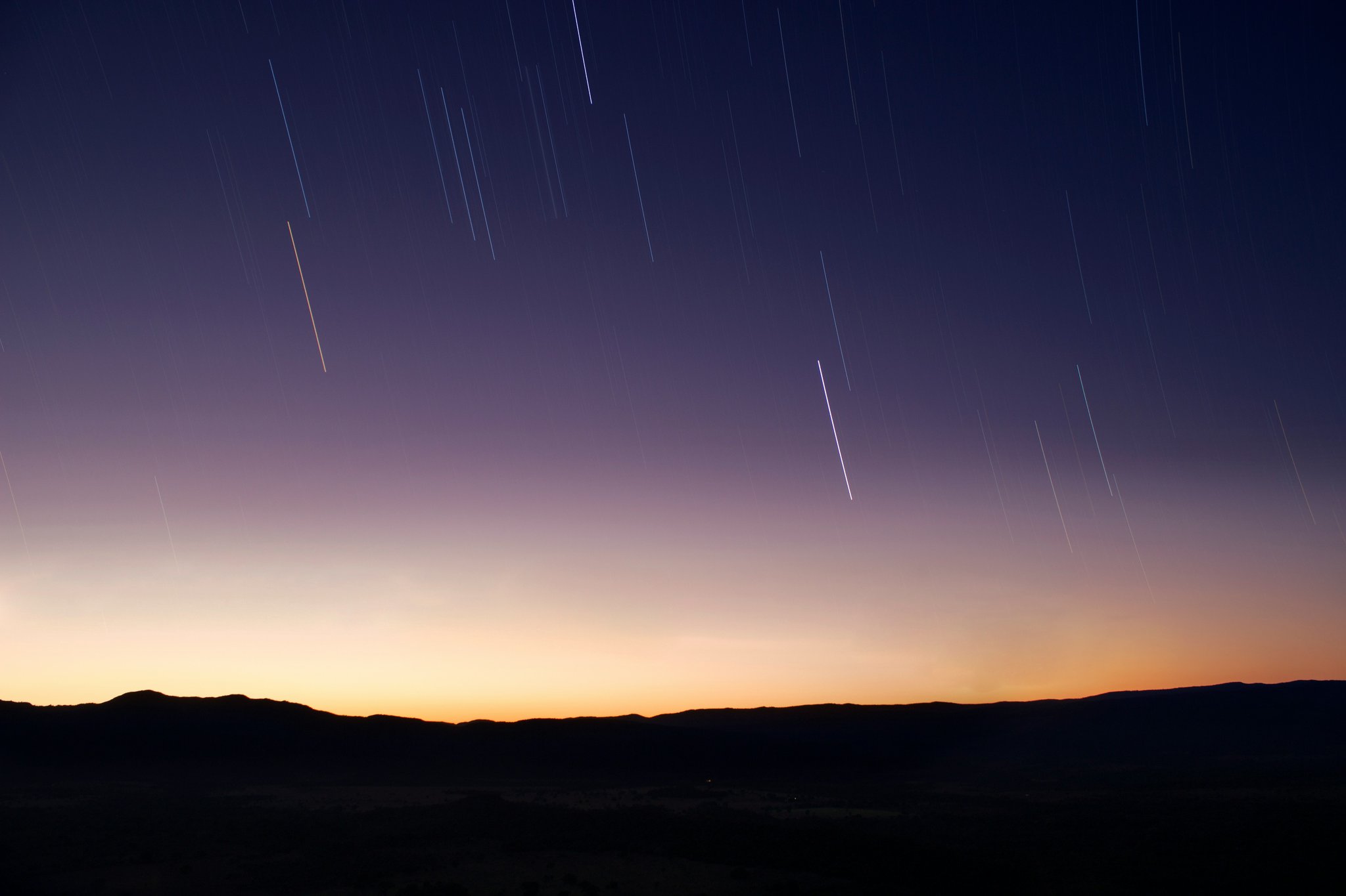 Rastros de estrelas em longa exposição cruzam um céu crepuscular roxo-escuro sobre as colinas silhuetadas da Chapada dos Veadeiros, com um horizonte dourado e alaranjado