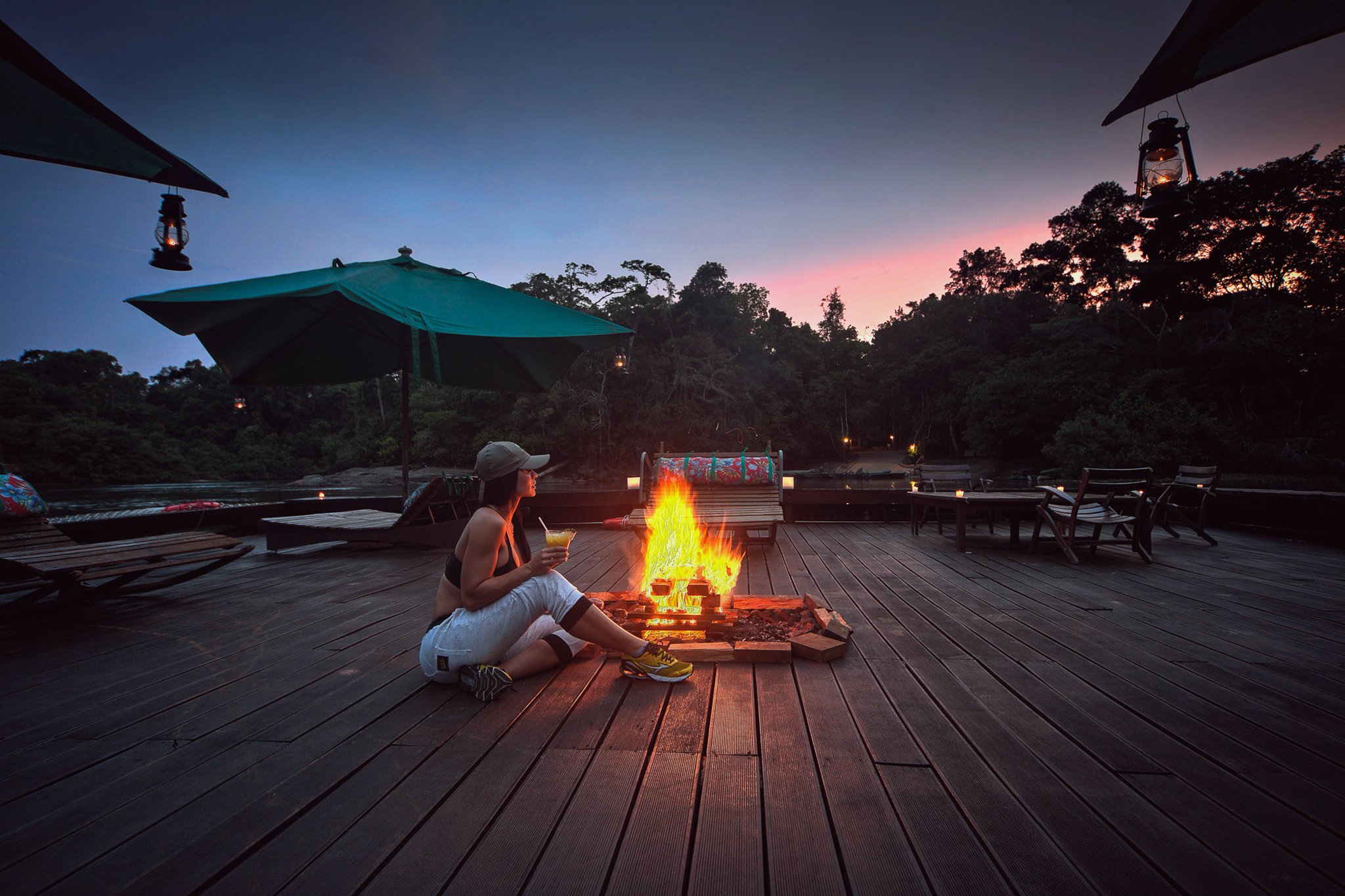 Cocktail in hand, fire crackling, jungle at the edge of the light. Photo: Samuel Melim A guest sits beside an open fire on the wooden deck at Cristalino Jungle Lodge, holding a cocktail as a pink-and-blue twilight sky glows above the rainforest canopy, with hanging lanterns lighting the scene