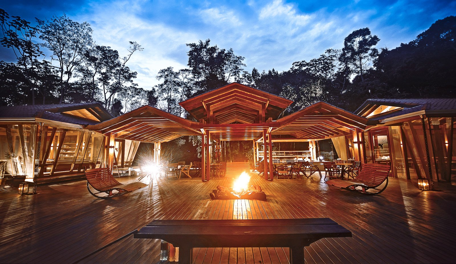 Firelight on polished hardwood — the lodge's communal deck after dark. Photo: Samuel Melim (c) The open-air timber deck at Cristalino Jungle Lodge glows at dusk, with a crackling bonfire at its centre, rocking chairs on either side, a bar visible in the background, and tall rainforest trees silhouetted against a deep blue twilight sky