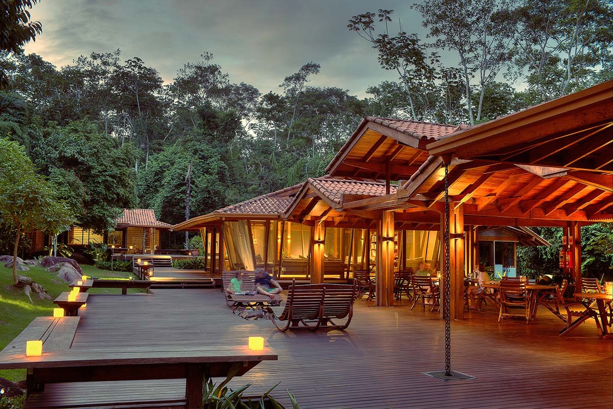 Glowing lanterns, rocking chairs, and the jungle breathing just beyond the railings. Photo: Samuel Melim Cristalino Jungle Lodge's timber open-air deck glowing with candles and warm lamp light at dusk, with guests relaxing on wooden rocking chairs and dense rainforest rising in the background