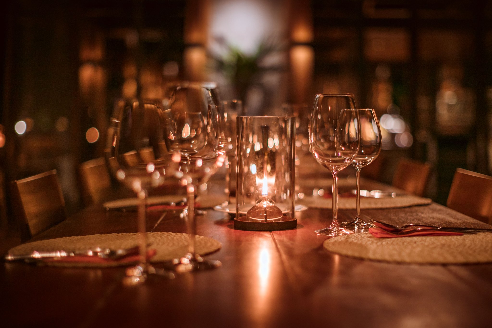 Candlelight, crystal, and the quiet hush of the forest just beyond the walls. Photo: Samuel Melim A long polished wooden dining table set for guests at Cristalino Jungle Lodge, with crystal wine glasses, woven placemats, red napkins, and a glowing glass candle lantern casting warm amber light across the room
