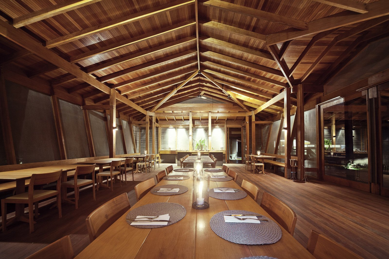Sustainably built from local hardwood, the dining room gathers guests under a cathedral ceiling each evening. Photo: Samuel Melim (c) The Cristalino Jungle Lodge restaurant dining room with a soaring vaulted timber ceiling, long communal wooden tables set with woven placemats and candles, and warm pendant lighting