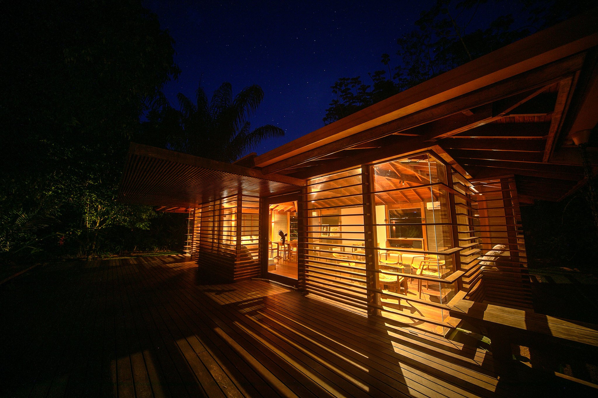 Warm light spills across the deck long after the jungle falls quiet. Photo: Samuel Melim Cristalino Jungle Lodge glowing from within at night, its timber-slatted walls and wide wooden deck illuminated by warm amber light beneath a star-filled sky