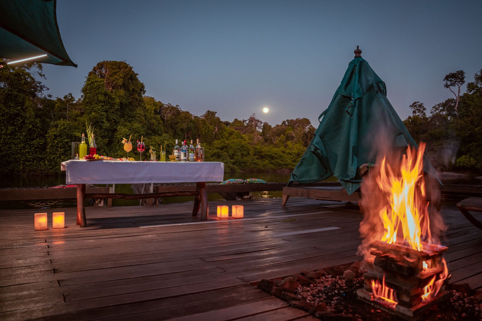 Cocktails, candlelight, and a crackling bonfire as the full moon climbs over the forest. Photo: Samuel Melim An open timber deck at Cristalino Jungle Lodge at dusk, with a roaring bonfire in the foreground, paper lantern candles lining the floor, a white-clothed cocktail table set with colorful drinks and bottles, and a full moon rising above the rainforest treeline