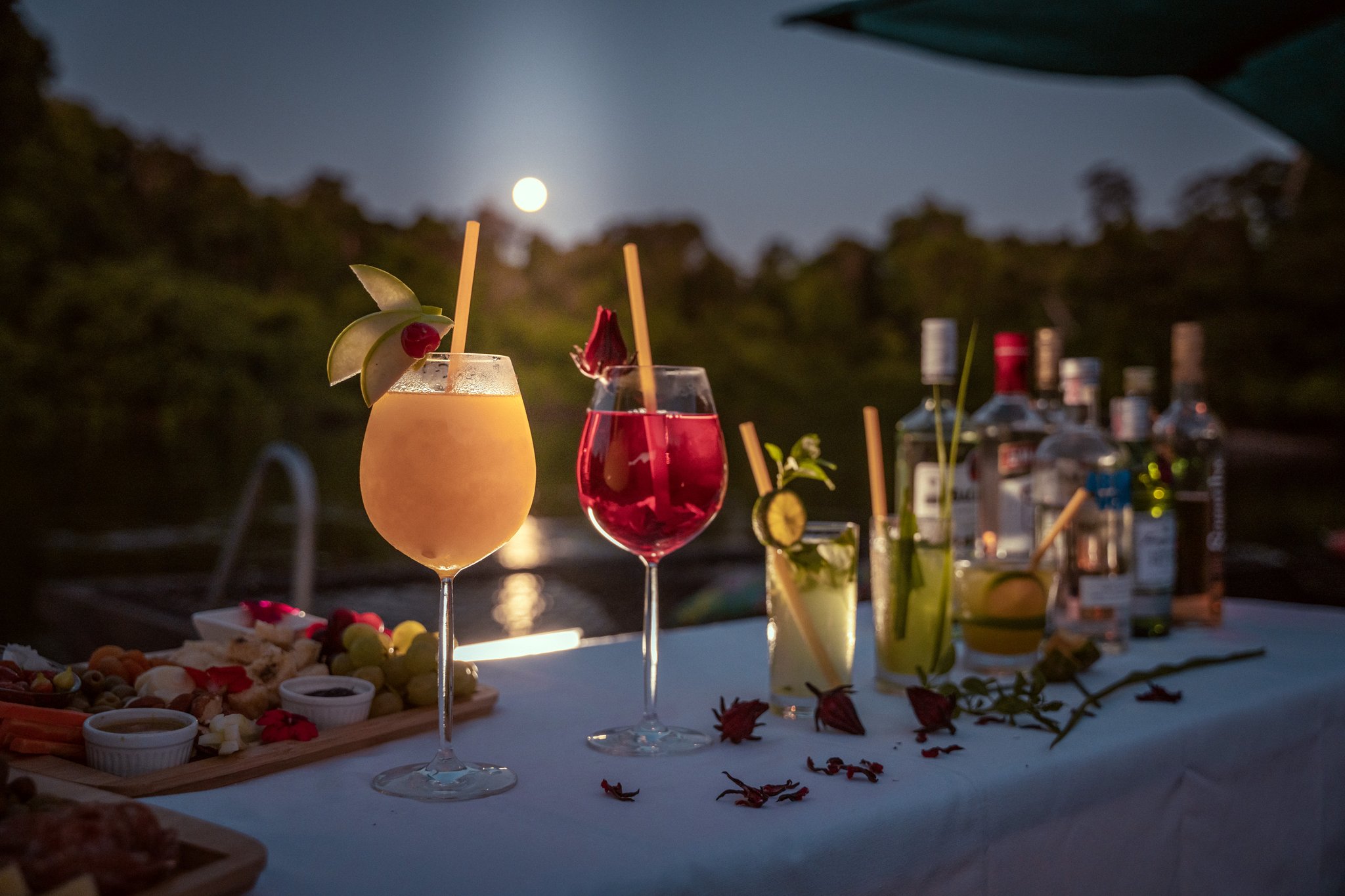 Full moon, four cocktails, and the jungle humming just beyond the table. Four garnished cocktails and a charcuterie board on a white-draped table at Cristalino Jungle Lodge, with a full moon rising over the rainforest canopy in the background