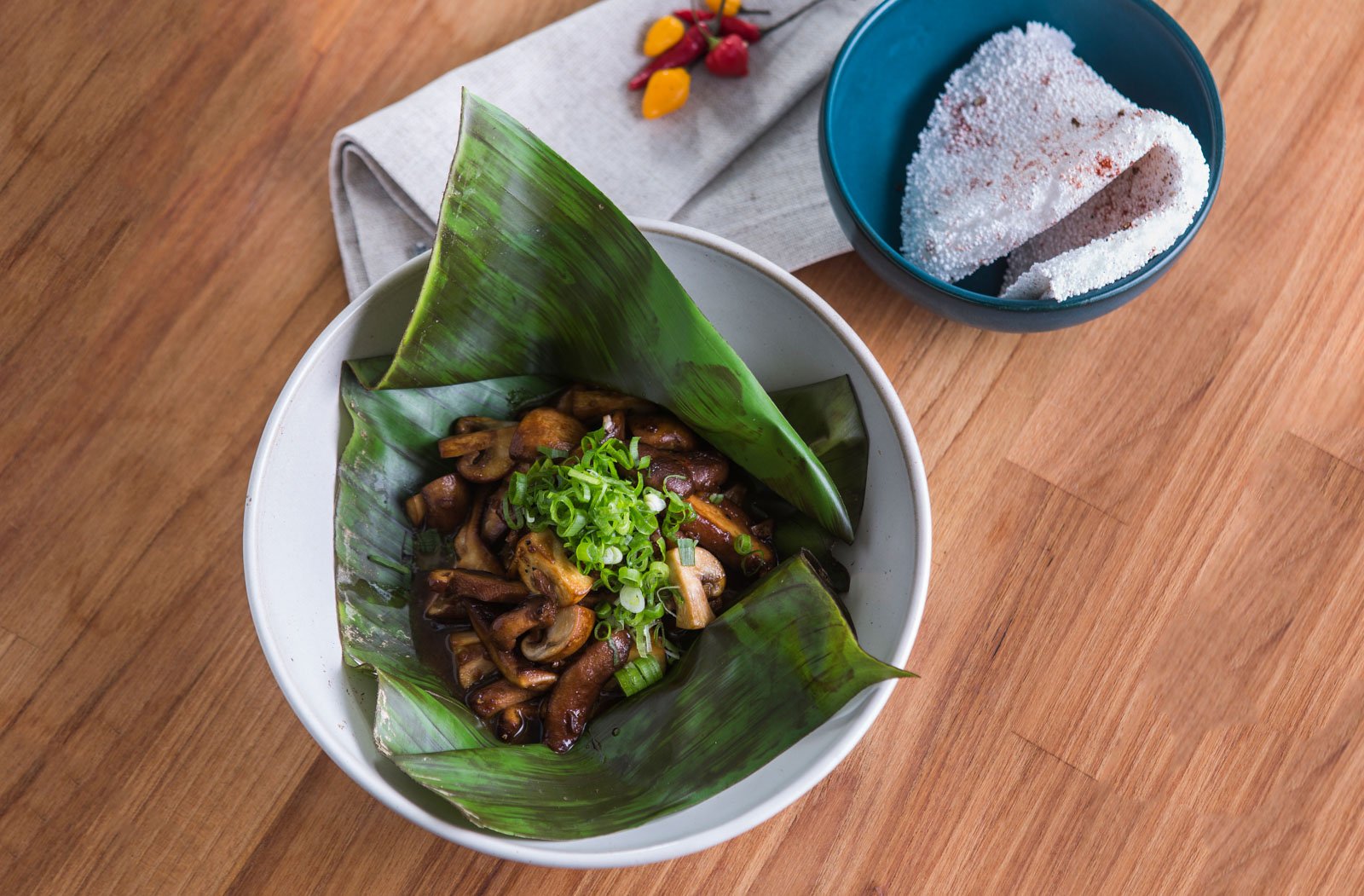 Mushrooms, banana leaf, a scatter of spring onion — forest cooking at its most honest. Photo: Rjcastilho Sautéed mushrooms garnished with sliced spring onions, served in a banana leaf inside a white ceramic bowl at Cristalino Jungle Lodge, with a side of cassava flatbread dusted in red spice and small chilli peppers on a linen napkin