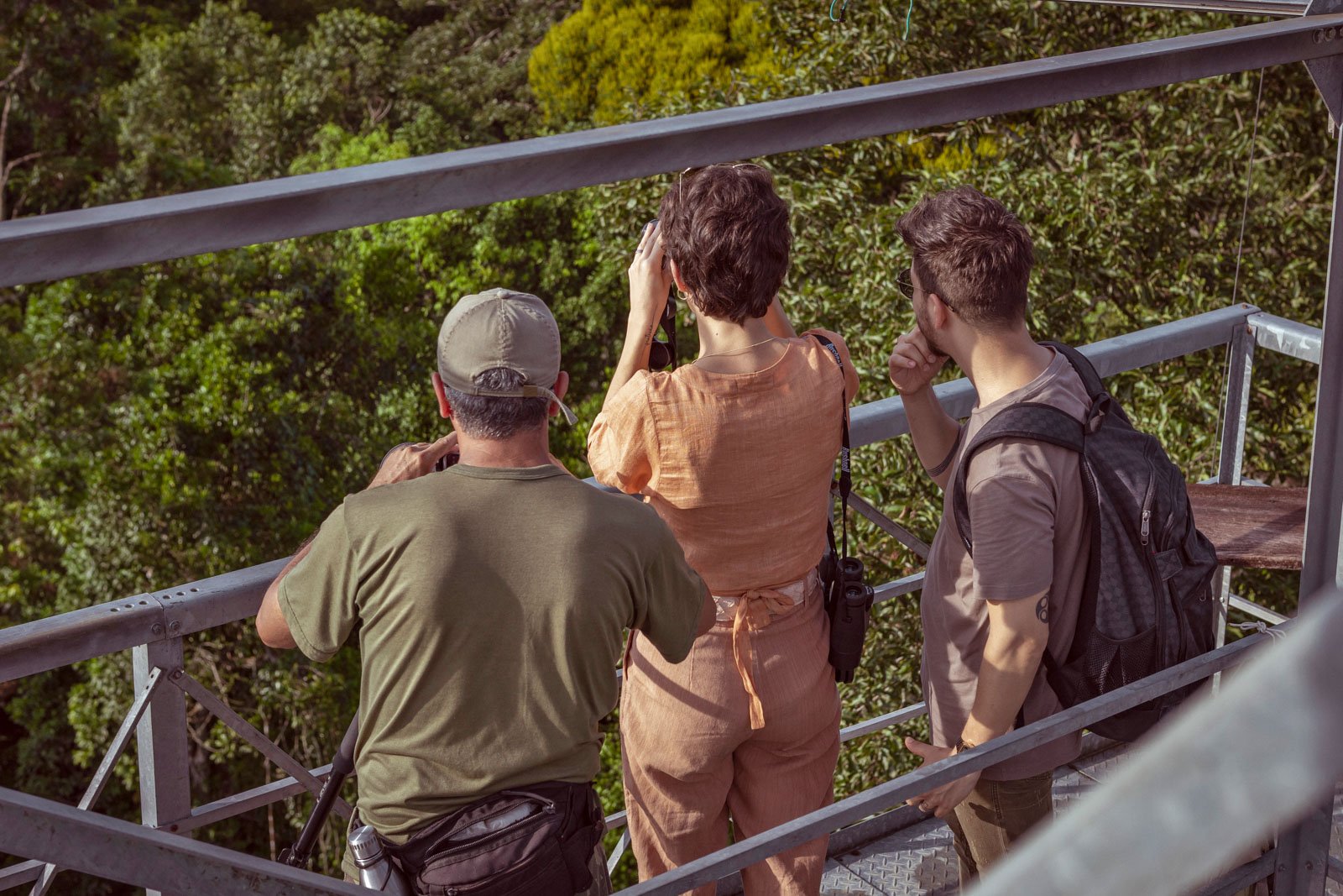 Binoculars up, breath held — the canopy always has something to reveal. Photo: Samuel Melim Three visitors scan the rainforest canopy with binoculars from a steel observation tower at Cristalino Jungle Lodge