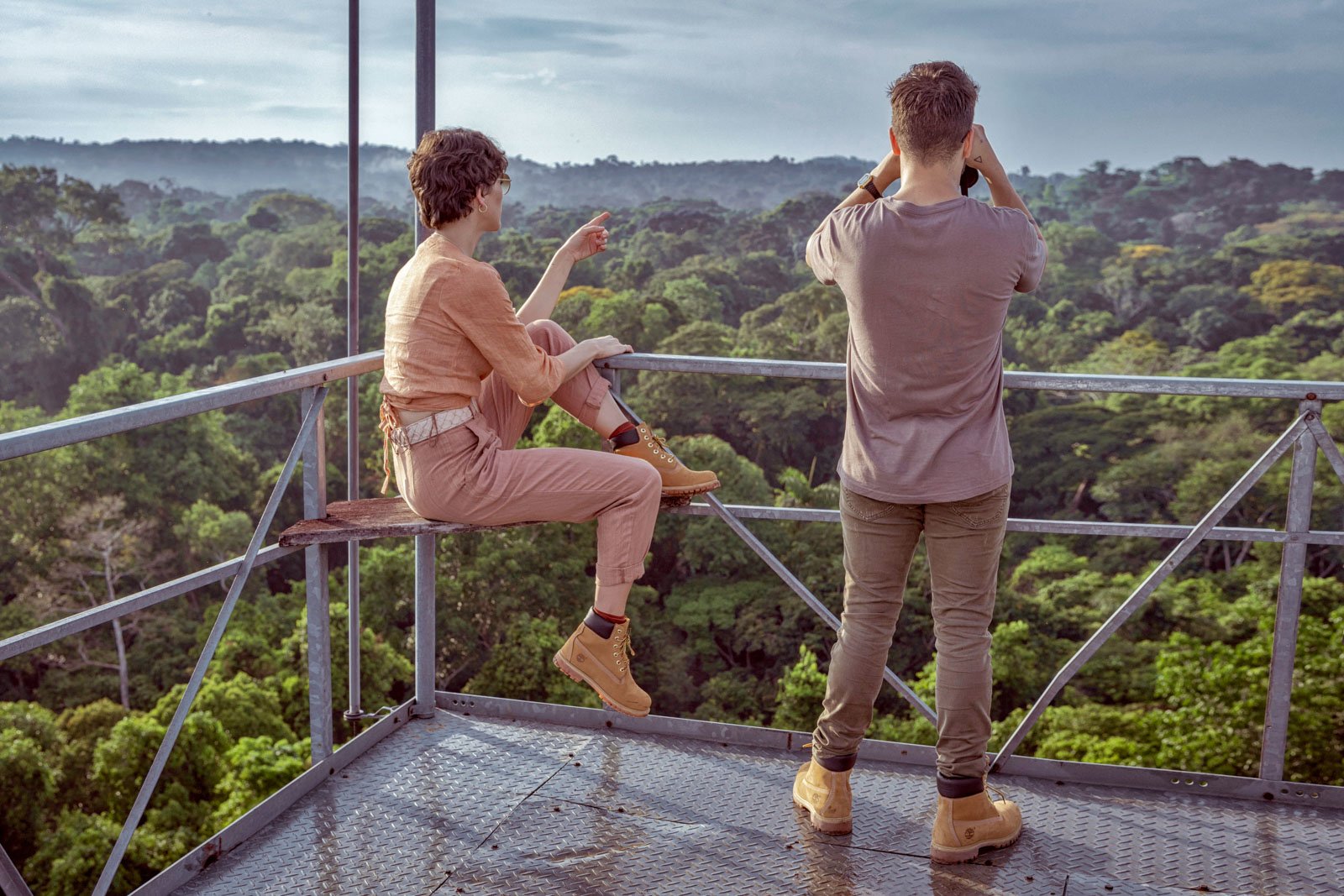 Two travellers on a metal observation tower platform at Cristalino Jungle Lodge, one seated pointing across an endless green rainforest canopy, the other scanning with binoculars under a misty sky