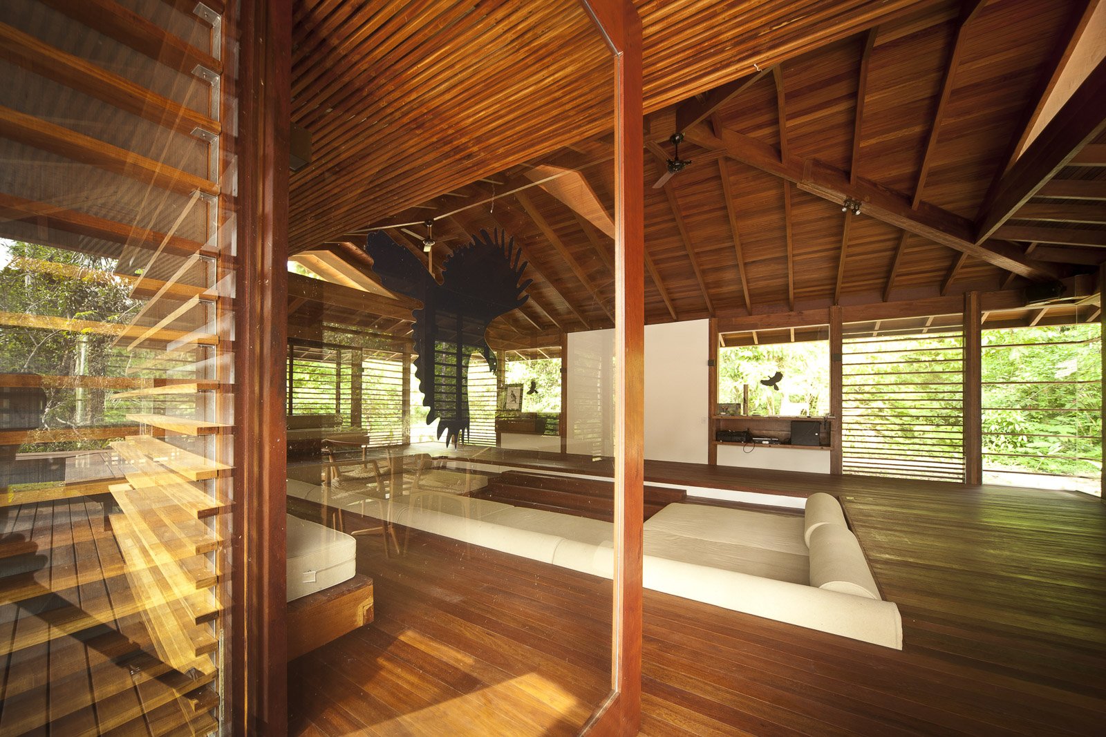 Rich timber, white cushions, and a canopy of green just beyond the louvres. Photo: Samuel Melim (c) Open-air common lounge at Cristalino Jungle Lodge featuring warm hardwood floors, a vaulted timber ceiling, white day-bed sofas, and louvred walls opening onto lush green rainforest