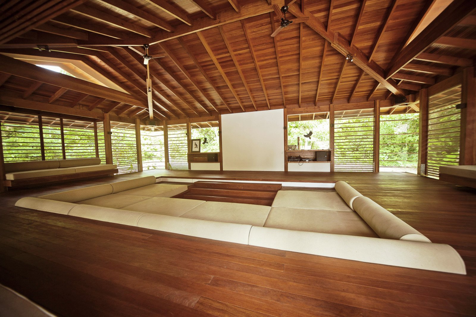 A cinema under the canopy — cream cushions, tropical timber, and the jungle just beyond the louvres. Photo: Samuel Melim (c) The open-air lounge at Cristalino Jungle Lodge features a sunken seating area with cream cushions, polished hardwood floors, a large projector screen, and a vaulted timber roof with ceiling fans, surrounded by lush green forest on all sides.
