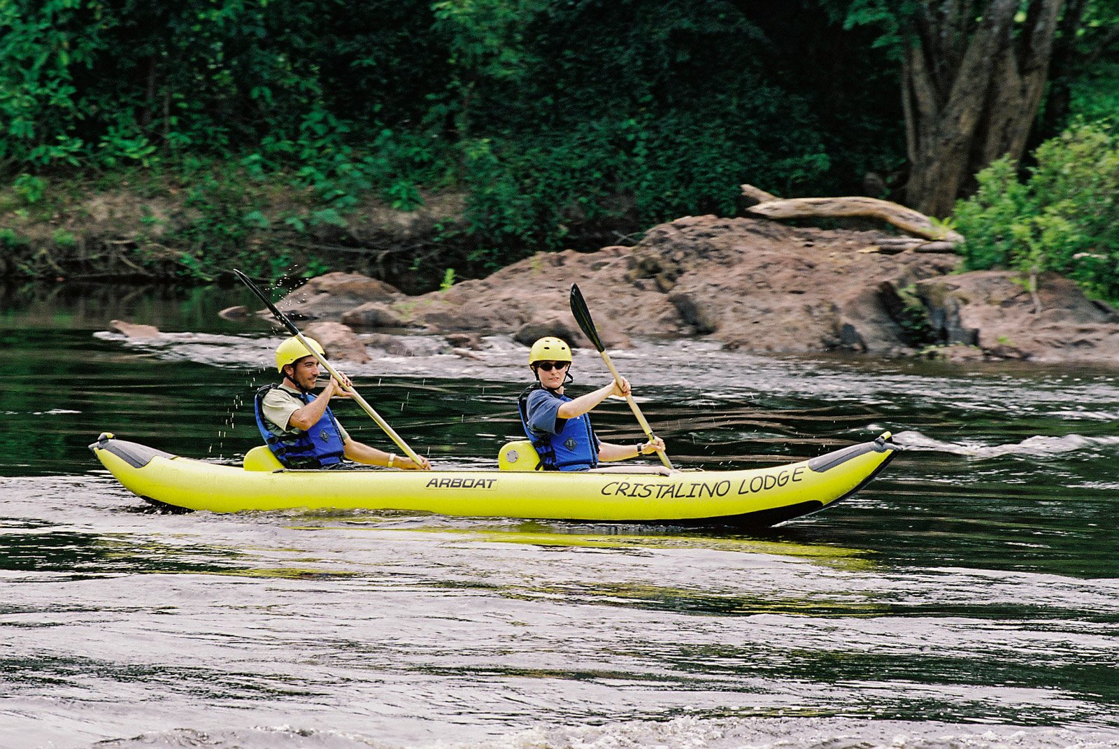 Two guests in yellow helmets and life jackets paddling a yellow Cristalino Jungle Lodge inflatable tandem kayak along a calm jungle river with forested rocky banks