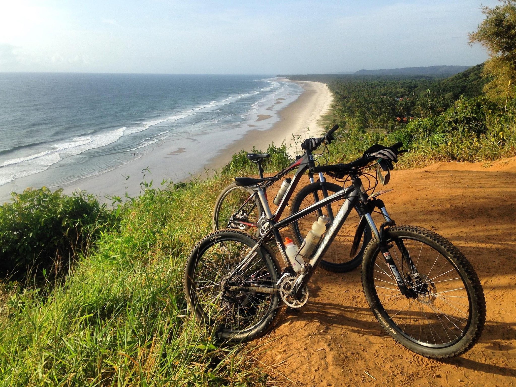 The reward for every climb — endless coastline stretching south from Serra Grande. Two mountain bikes resting on a red dirt track during a cycling Bahia journey, with a sweeping panorama of a long white-sand beach, rolling Atlantic waves and dense green Atlantic Forest stretching to the horizon