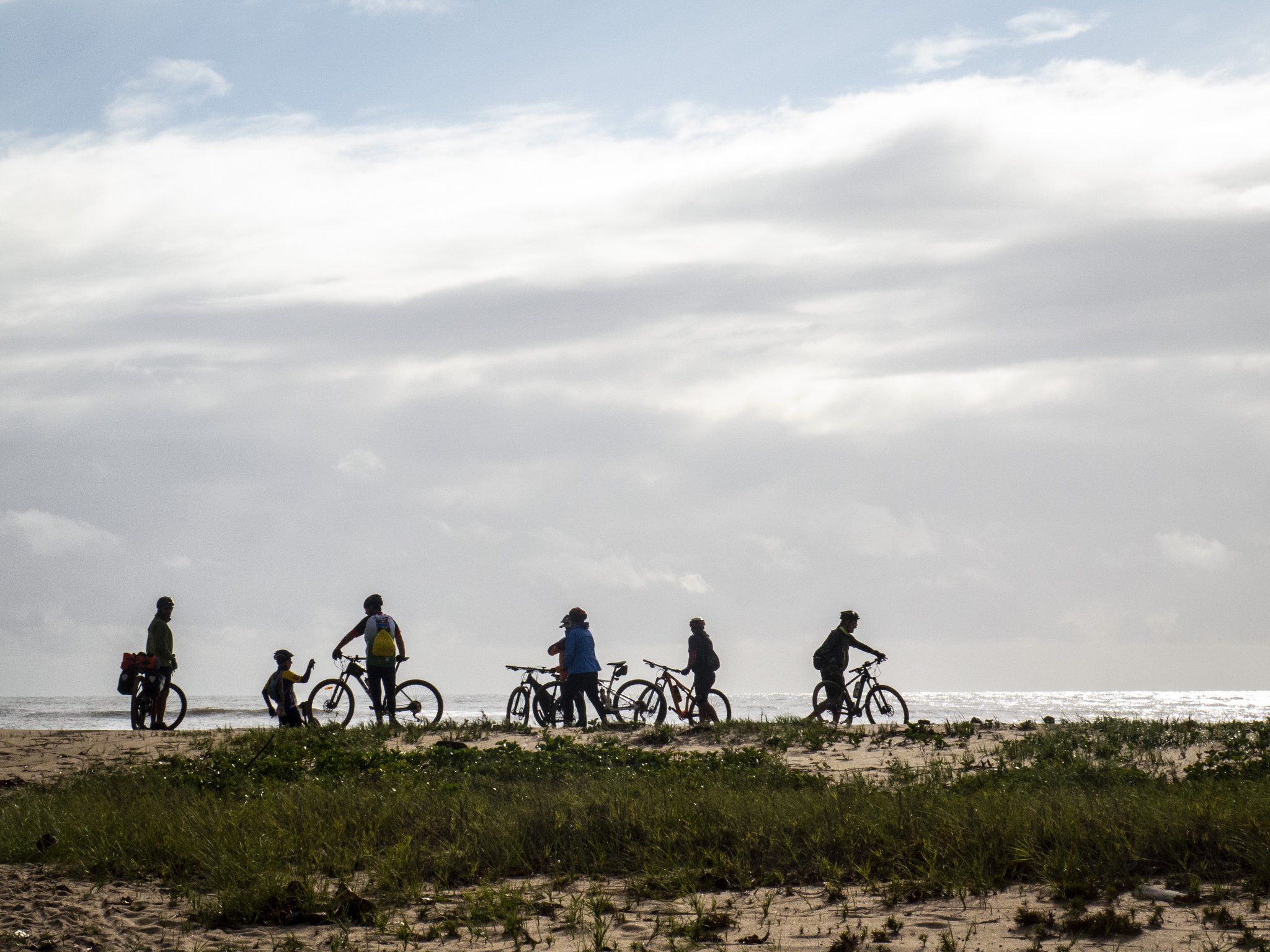 A brief pause on the sands — the coast of Bahia stretches endlessly ahead. Silhouettes of five cyclists with mountain bikes pausing on a sandy coastal dune with low vegetation and the Atlantic Ocean glimmering behind them under a dramatic cloudy sky — a scene from a cycling Bahia itinerary between Ilhéus and Itacaré