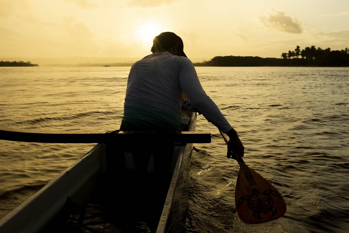 Golden hour on the Rio das Contas — paddling into the Bahian sunset. A paddler silhouetted against a golden sunset steers a traditional Hawaiian canoe across the calm Rio das Contas near Itacaré, on a cycling Bahia tour