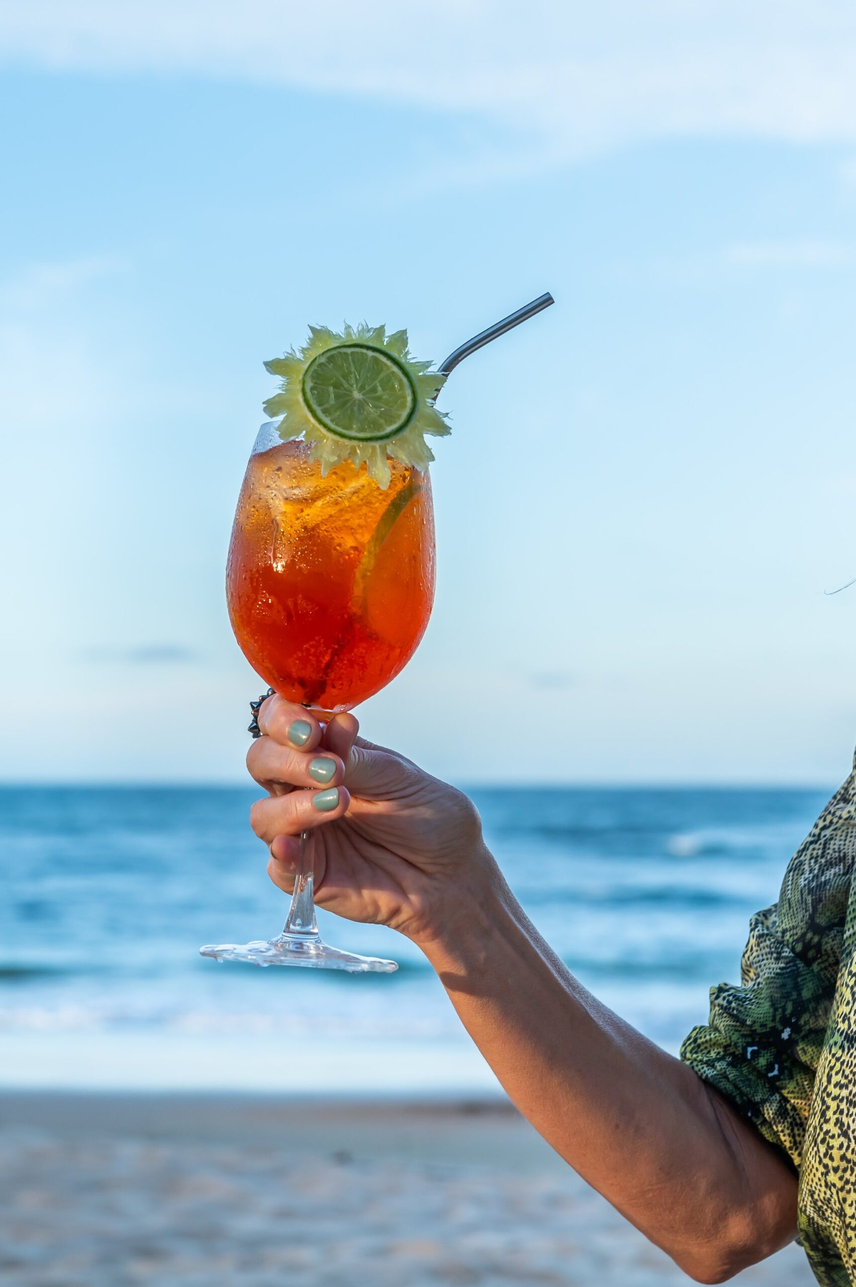 Sip something tropical — Itacaré's beachside refreshments are well earned. A hand holding an amber tropical cocktail garnished with lime and a metal straw, with the ocean and blue sky of Itacaré beach in the background during a cycling Bahia tour