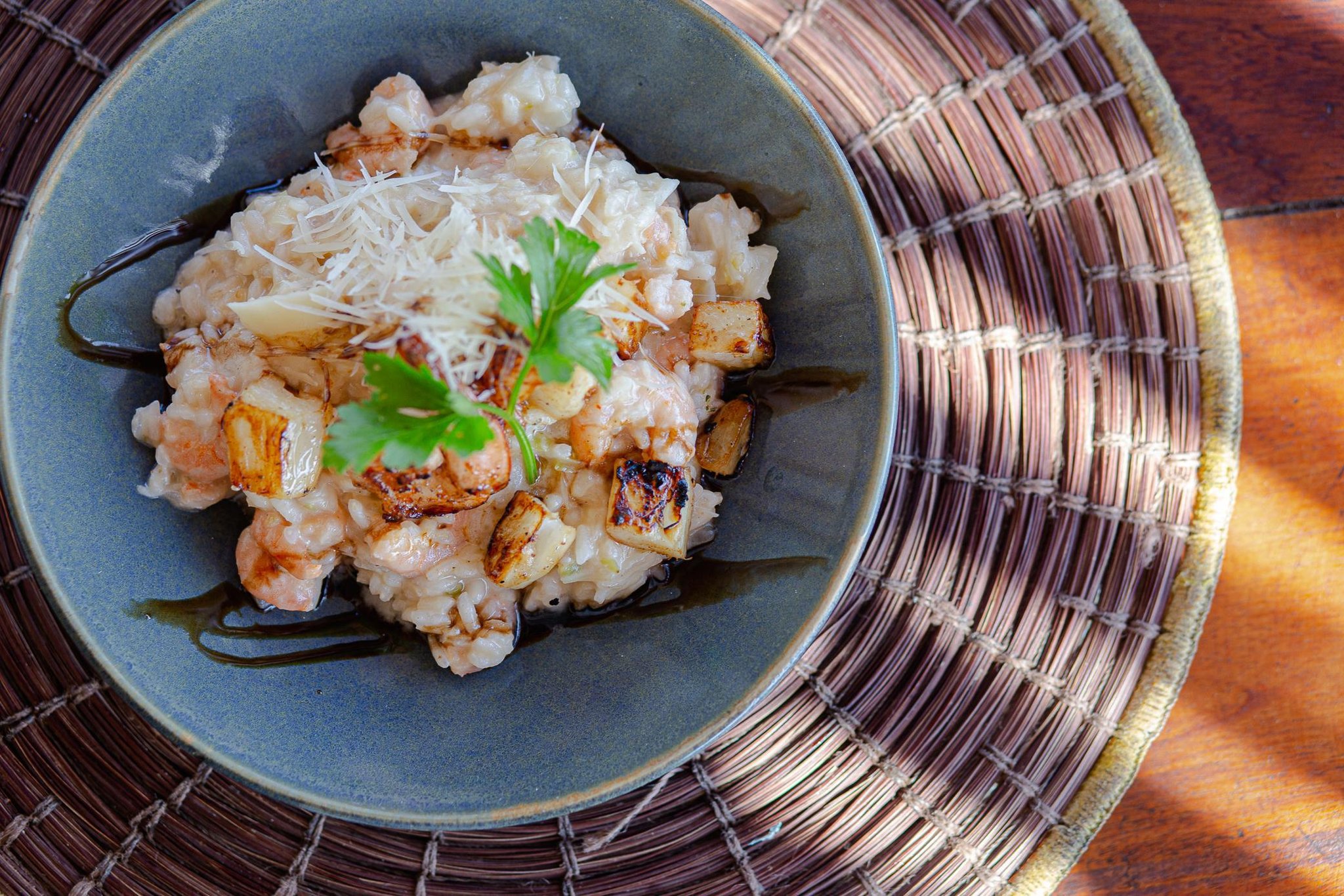 Farm-fresh flavours on the Cocoa Route — a beautifully plated risotto after a day in the saddle. Overhead view of a creamy shrimp and roasted vegetable risotto topped with grated parmesan and fresh parsley, served in a slate-blue ceramic bowl on a woven rattan placemat in Itacaré, Bahia