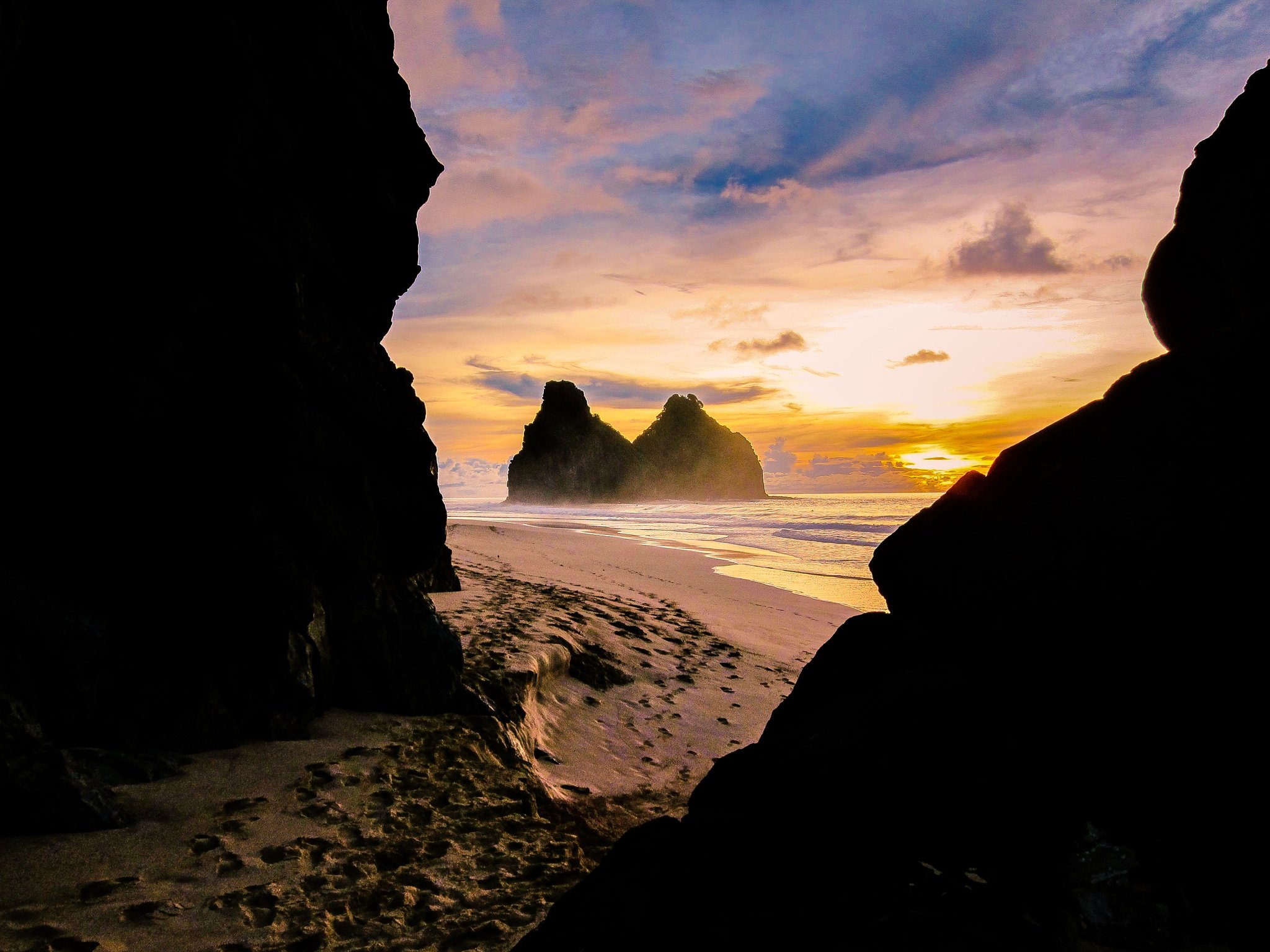 View through dark rock formations framing twin sea stack silhouettes and a golden sunset sky over the beach in Fernando de Noronha, a premier destination for fernando de noronha diving packages