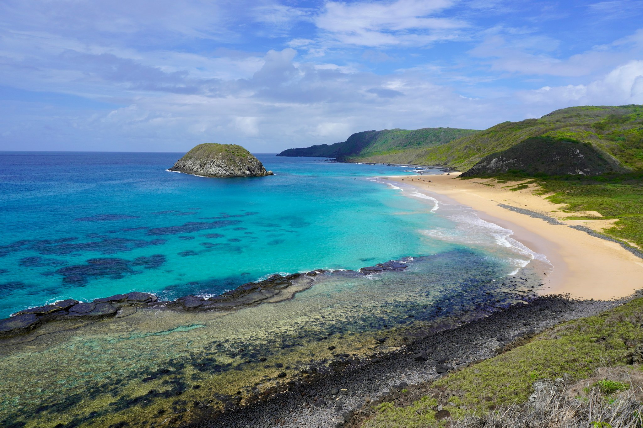 Vista aérea de uma praia em forma de crescente em Fernando de Noronha, com águas turquesas revelando formações de coral, plataformas de rocha vulcânica, um pequeno ilhote rochoso ao largo e colinas verdes emoldurando a baía