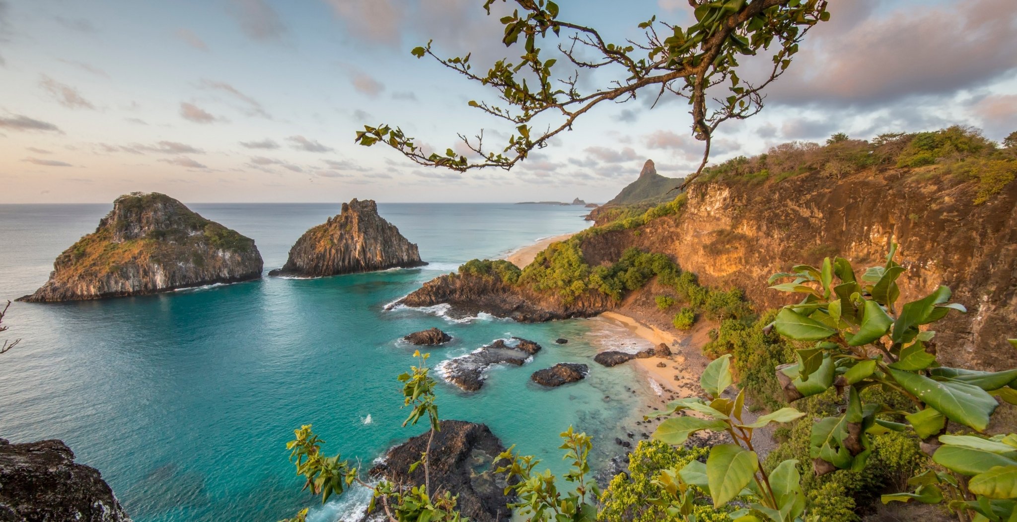 Panoramic clifftop view of a Fernando de Noronha diving trip destination showing turquoise bays, rocky sea stacks, a secluded sandy beach, and the iconic spire peak rising in the background at golden hour