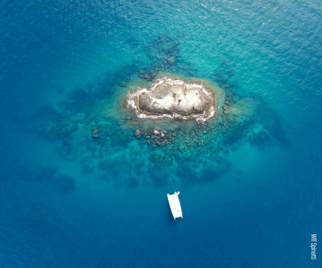 Vista aérea de um catamarã branco ancorado ao lado de um ilhote rochoso cercado por água turquesa cristalina em um mergulho em Fernando de Noronha