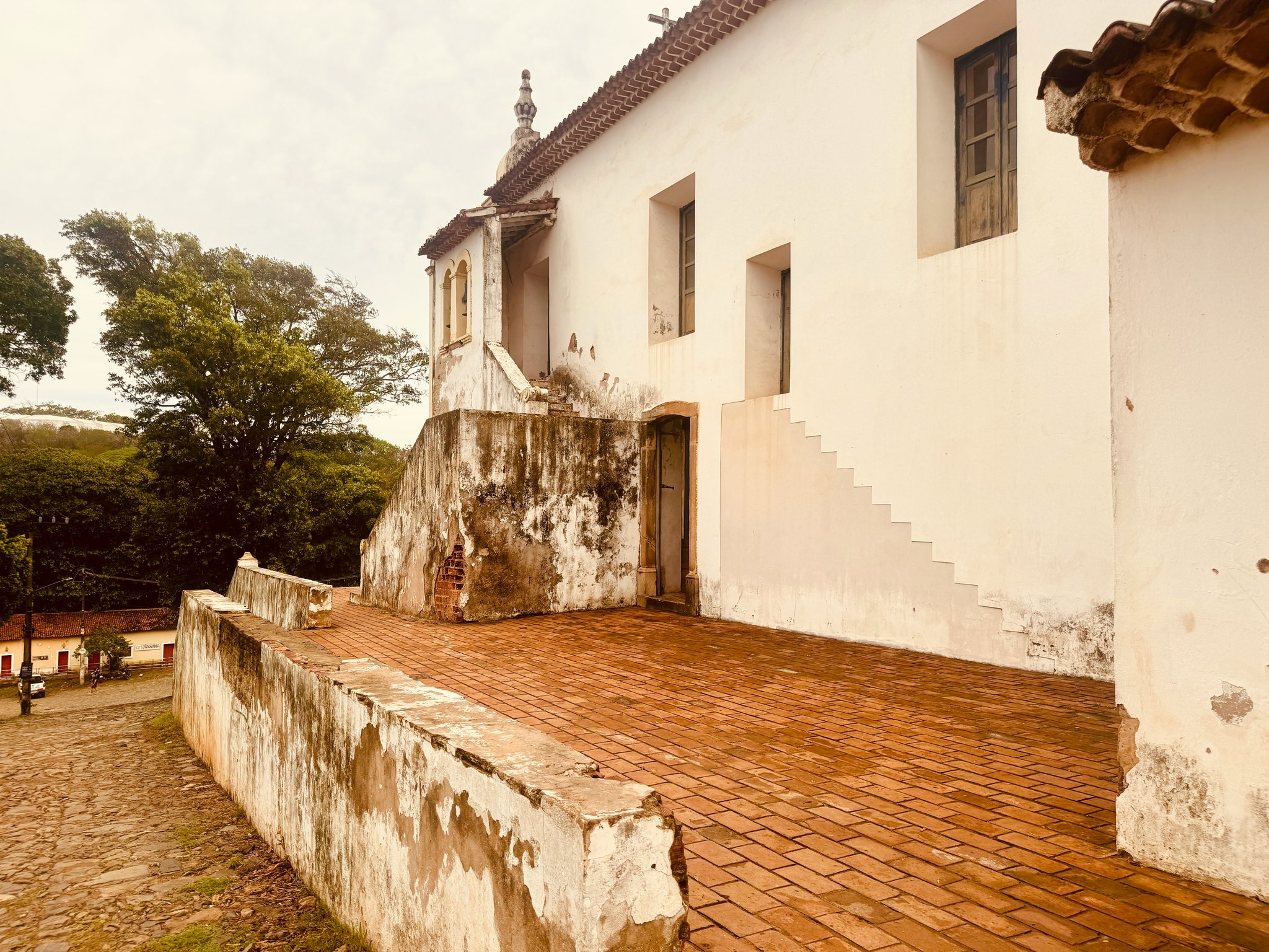 Fachada de igreja colonial branca com terraço de tijolos de barro e campanário com vista para uma praça de paralelepípedos em Fernando de Noronha