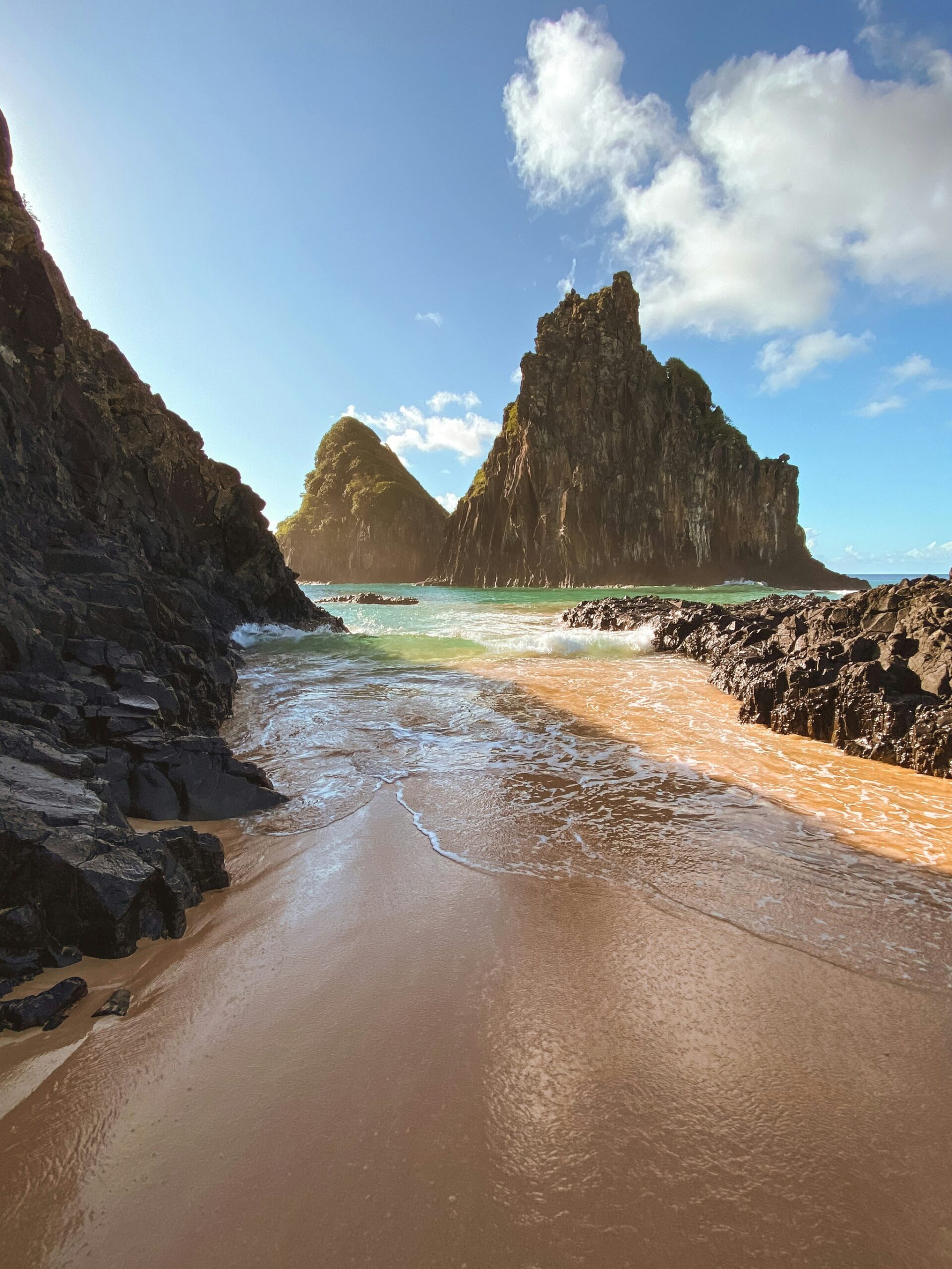 Uma enseada de areia dourada ladeada por rochas vulcânicas escuras se abre para as águas turquesa do Atlântico, com dois imponentes picos rochosos cobertos de vegetação ao fundo em Fernando de Noronha