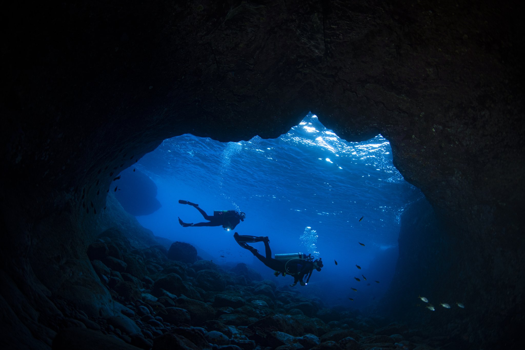 Dois mergulhadores em silhueta dentro de uma gruta subaquática rochosa com água azul elétrico em um mergulho em Fernando de Noronha, luz solar cintilando pela abertura da caverna