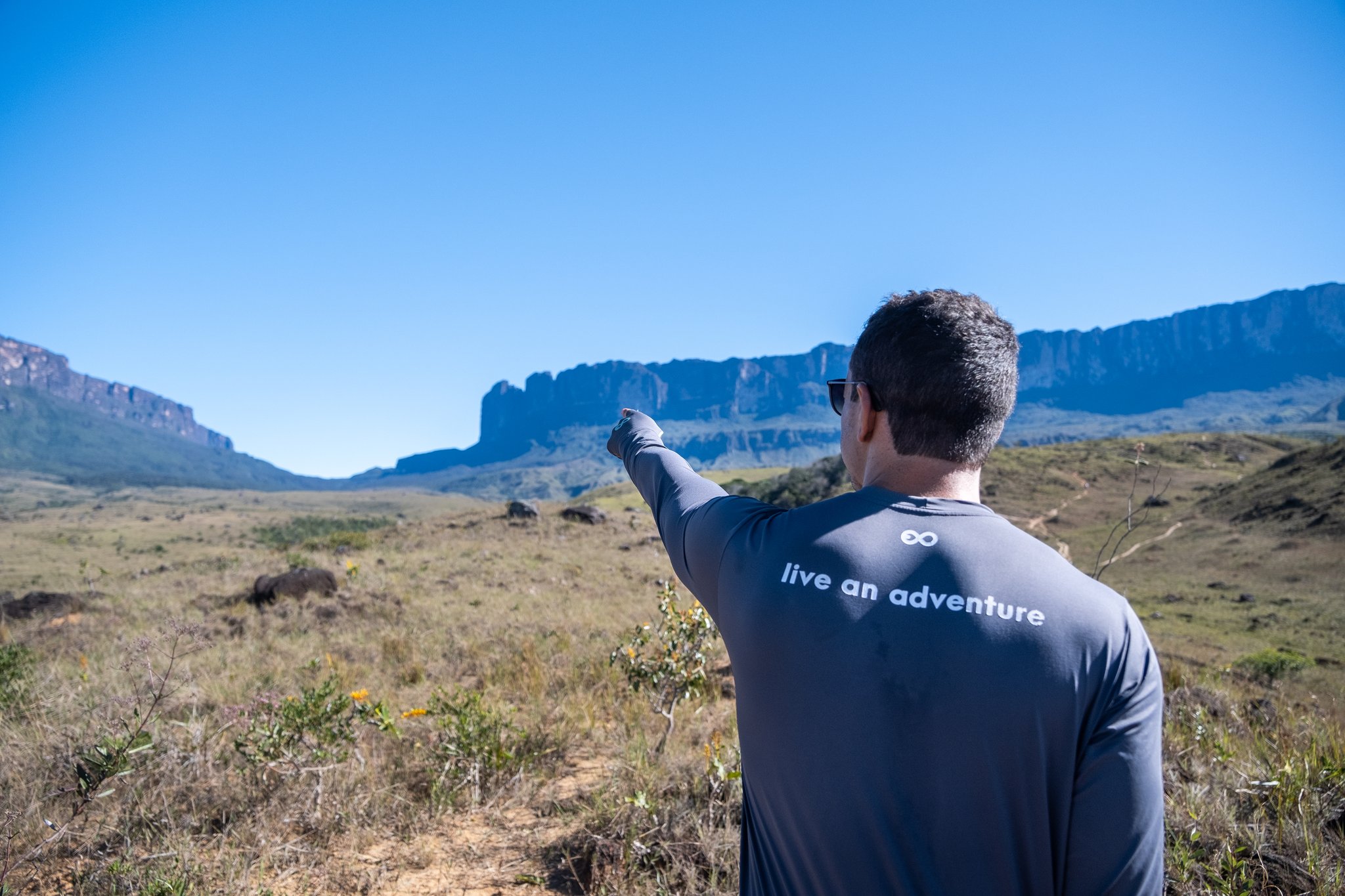 A hiker pointing toward the flat-topped cliffs of Mount Roraima across an open savanna plain, wearing a 'live an adventure' shirt — hiking in Brazil at its most dramatic