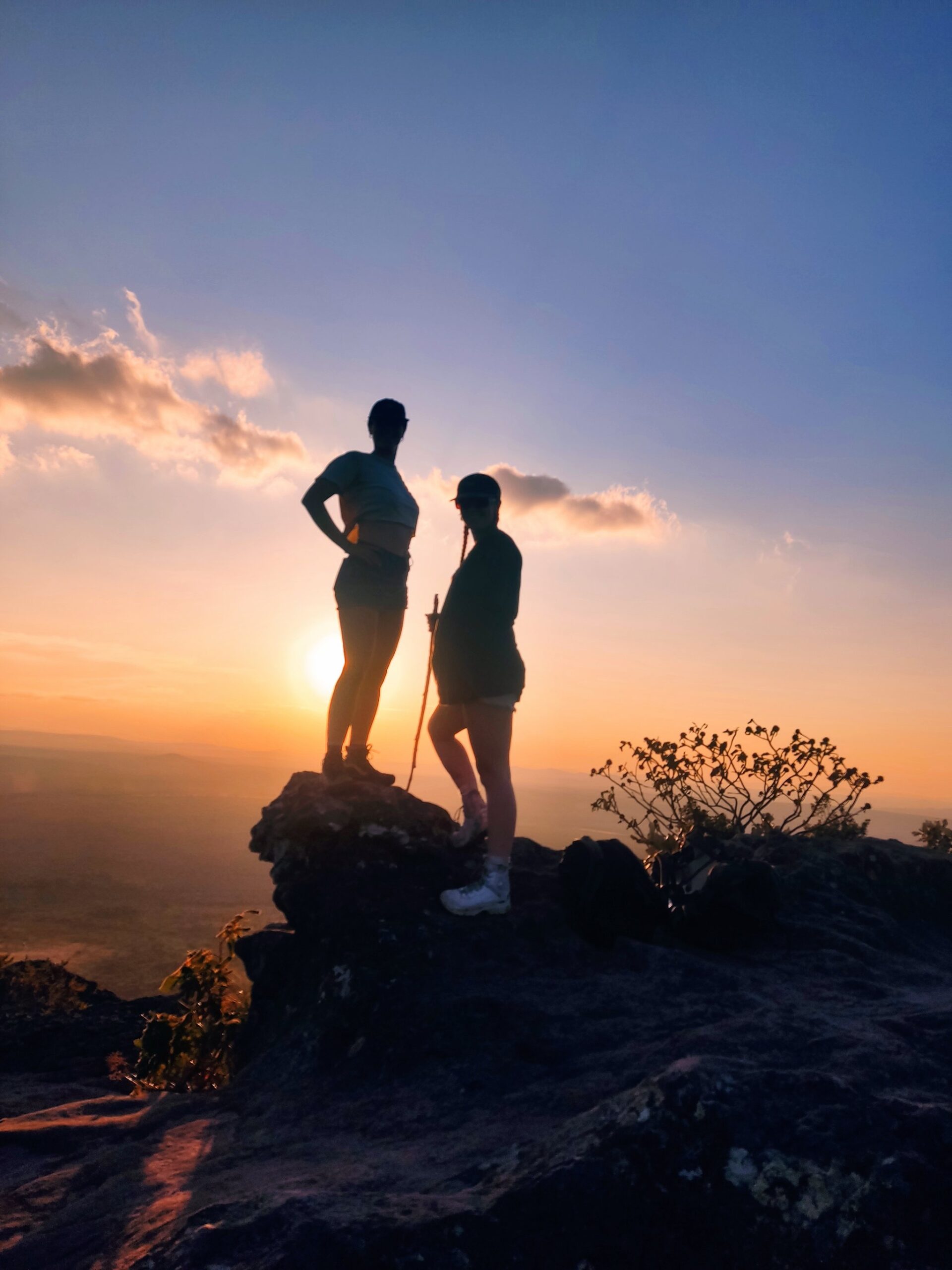Two hikers silhouetted against a vivid orange and blue sunset on a rocky summit during a hiking in Brazil adventure