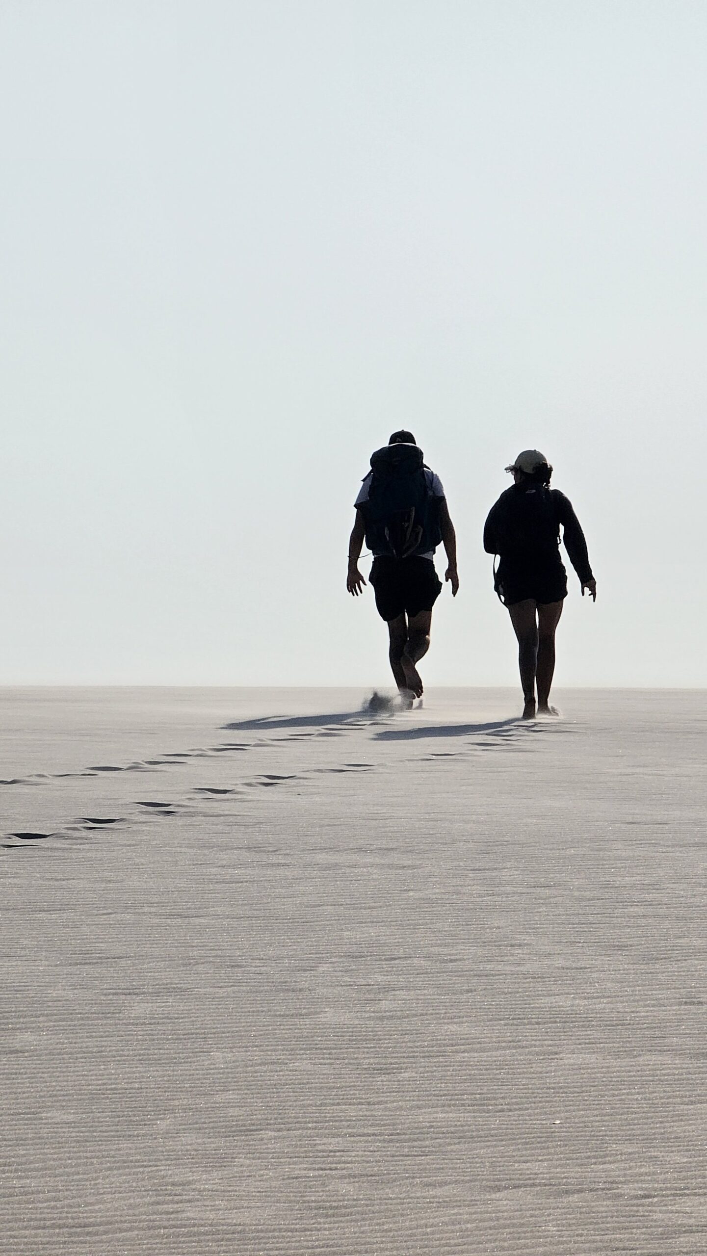 Two hikers with backpacks walking across a vast flat sand expanse during a hiking in brazil adventure, their footprints trailing behind them into the haze