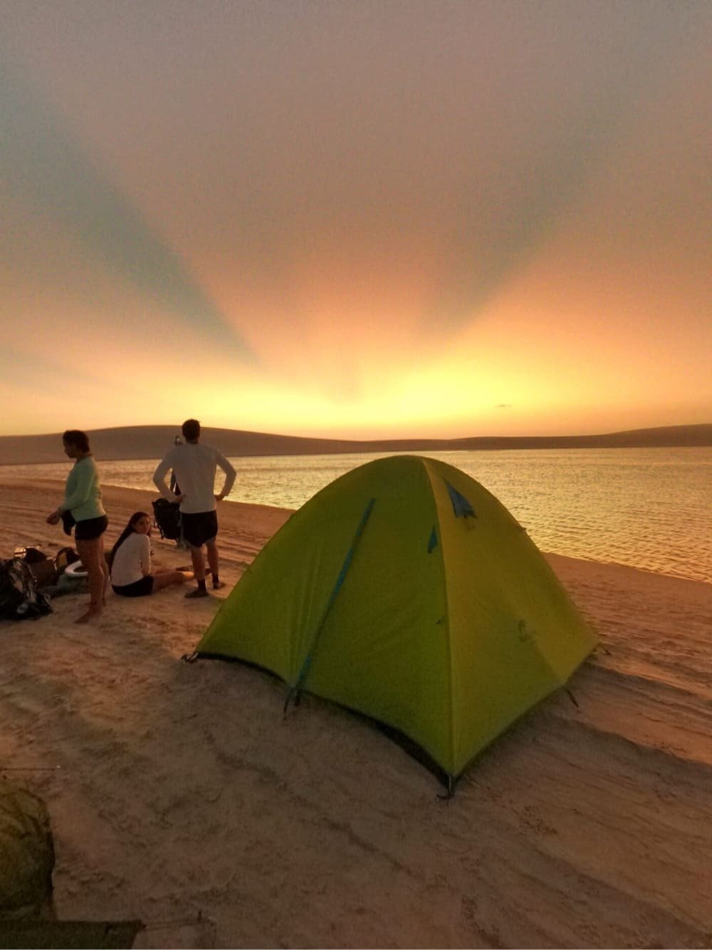 A lime-green dome tent pitched on a sandy riverbank at sunset during a horseback riding Lençóis Maranhenses tour, as three travellers watch golden rays radiate across a glassy waterway