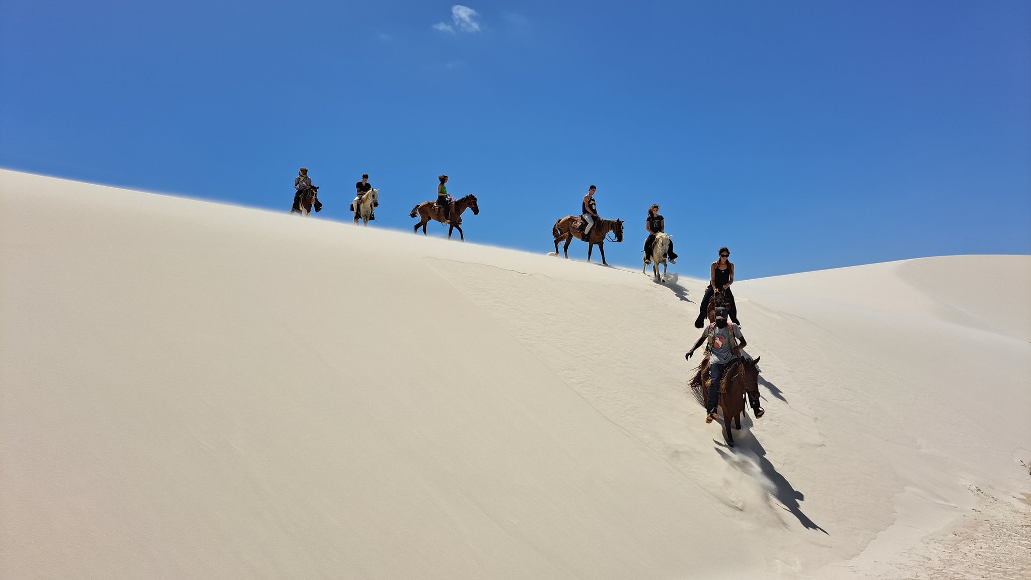 A line of riders on horseback descends a steep white sand dune crest during a horseback riding Lençóis Maranhenses tour, set against a vivid blue sky