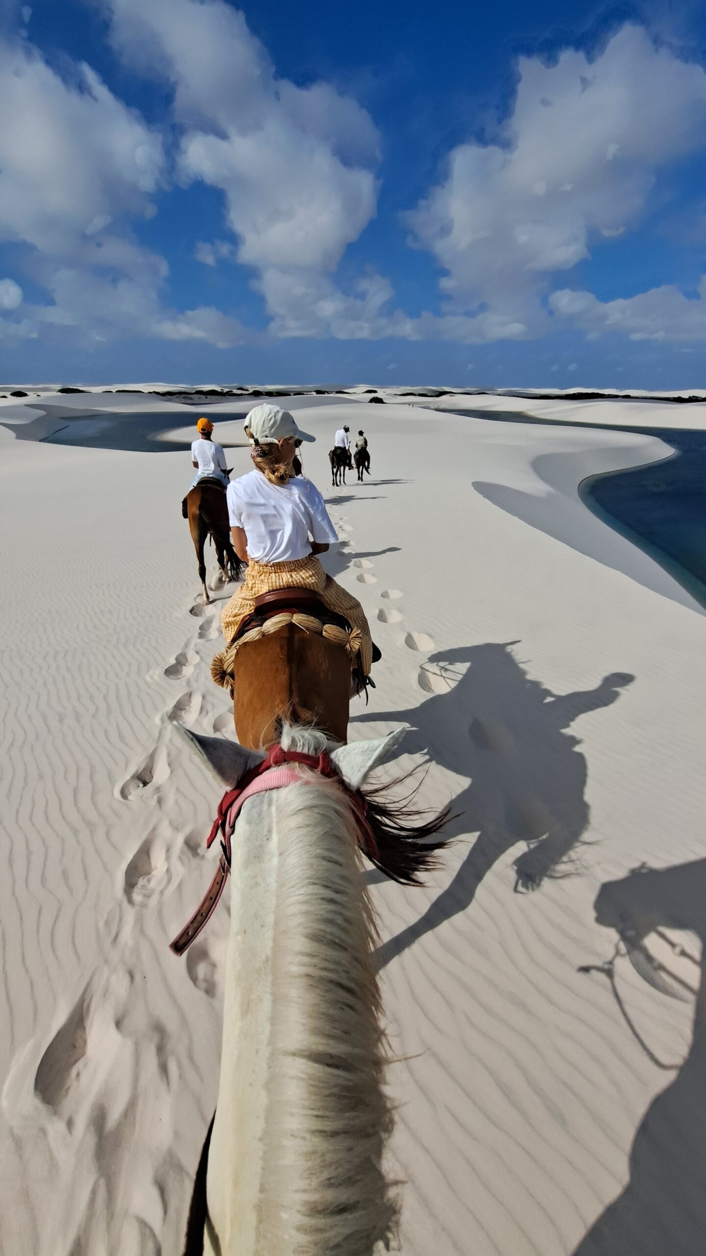 Point-of-view shot from horseback on a horseback riding Lençóis Maranhenses tour, with riders and hoofprints tracing a line across rippled white dunes beside a turquoise lagoon under a bright blue sky