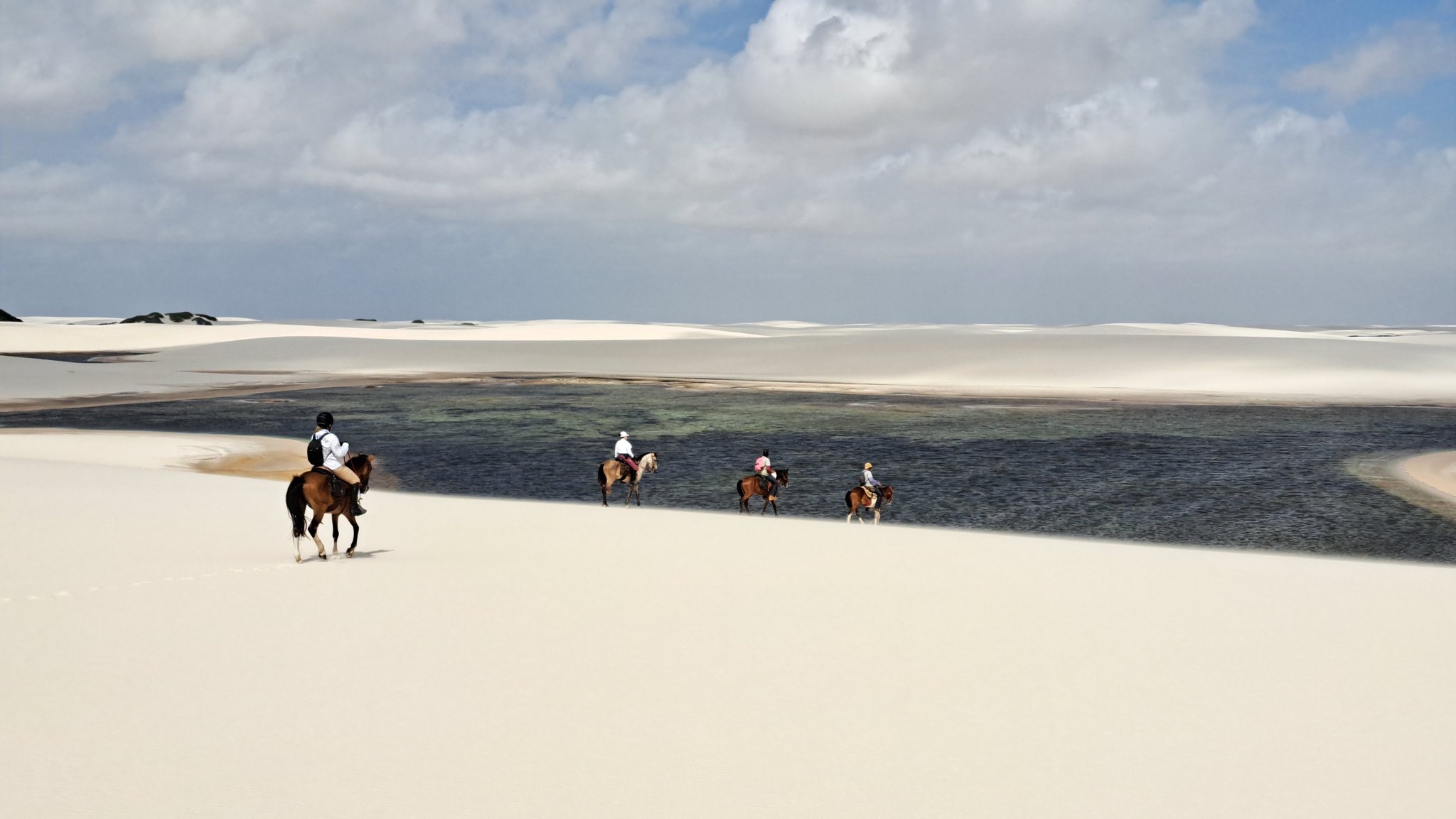 Four riders on horseback cross white sand dunes beside a dark freshwater lagoon on a horseback riding Lençóis Maranhenses tour, under a partly cloudy sky