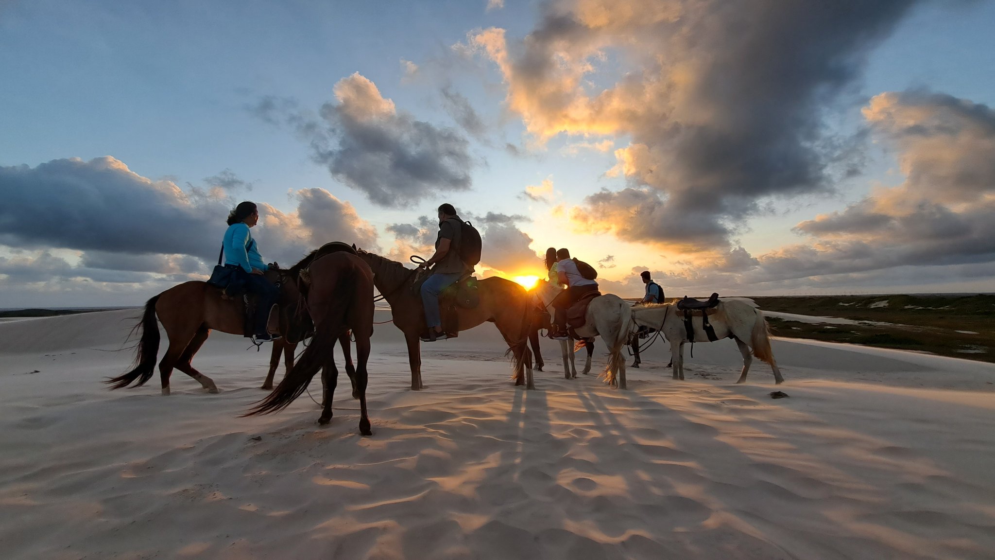 A group of riders on horseback silhouetted against a blazing sunset over the white sand dunes of Lençóis Maranhenses, with dramatic clouds spreading across the sky during a dune crossing on horseback