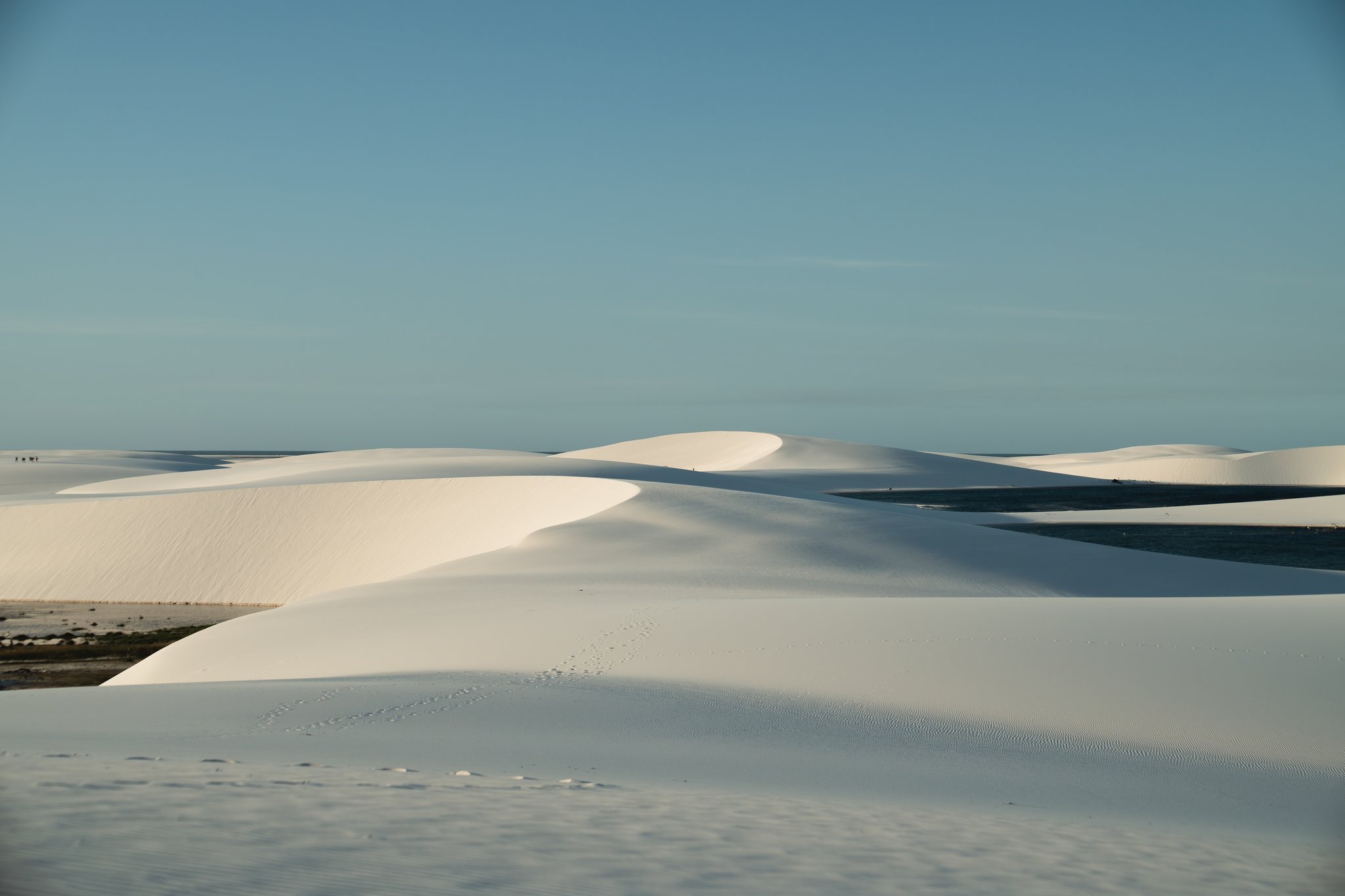 Sweeping white sand dunes of Lençóis Maranhenses stretch to the horizon beneath a clear blue sky, with shallow lagoons visible between the sculpted ridges on the horseback riding Lençóis Maranhenses tour route